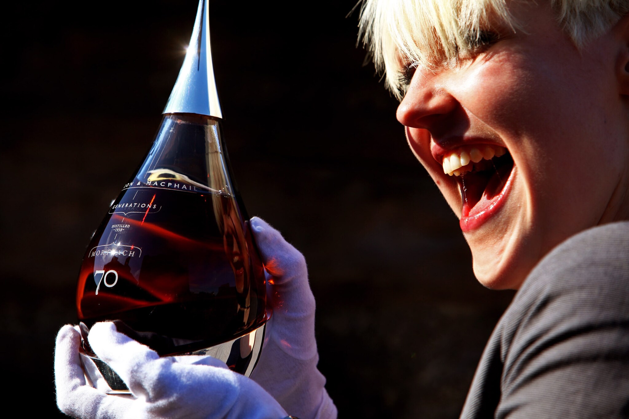 Eine lachende Frau hält mit weißen Baumwollhandschuhen eine tropfenförmige Flasche Whisky Eine lachende Frau hält mit weißen Baumwollhandschuhen eine tropfenförmige Flasche Whisky