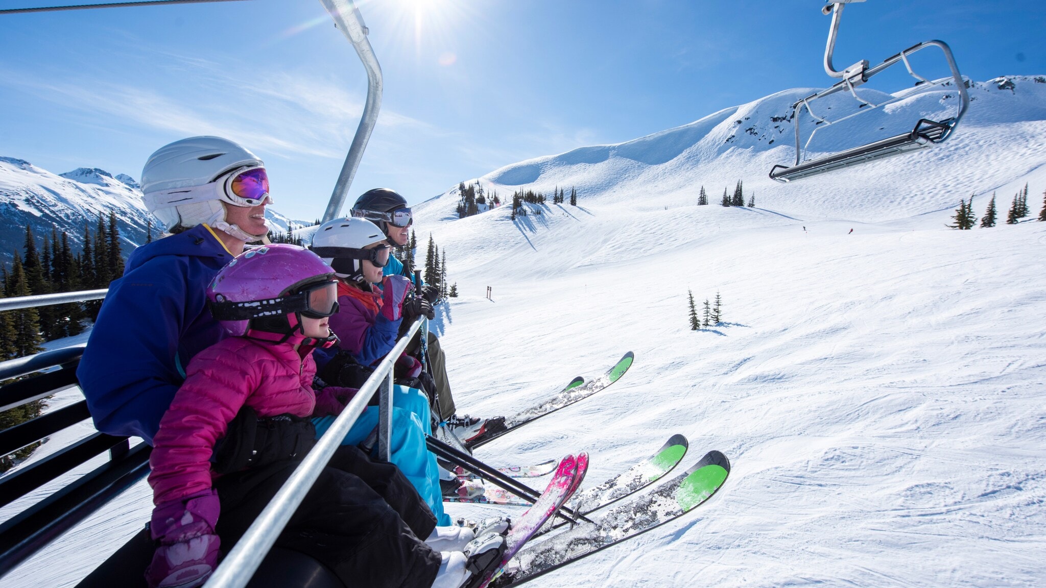 Eine Familie mit zwei Kindern auf Skiern sitzt in einem Sessellift über einer Skipiste bei Sonnenschein. Eine Familie mit zwei Kindern auf Skiern sitzt in einem Sessellift über einer Skipiste bei Sonnenschein.