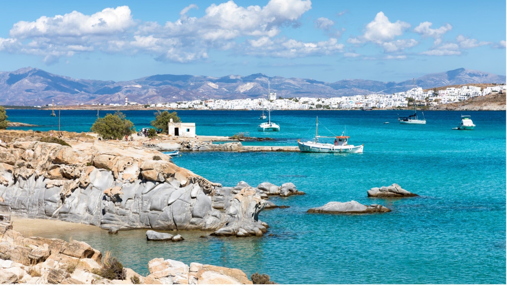 Blick auf eine felsige Küste mit klarem blauem Wasser, mehreren Segelbooten und einer weißen Stadt vor Bergen unter blauem Himmel mit Wolken Blick auf eine felsige Küste mit klarem blauem Wasser, mehreren Segelbooten und einer weißen Stadt vor Bergen unter blauem Himmel mit Wolken