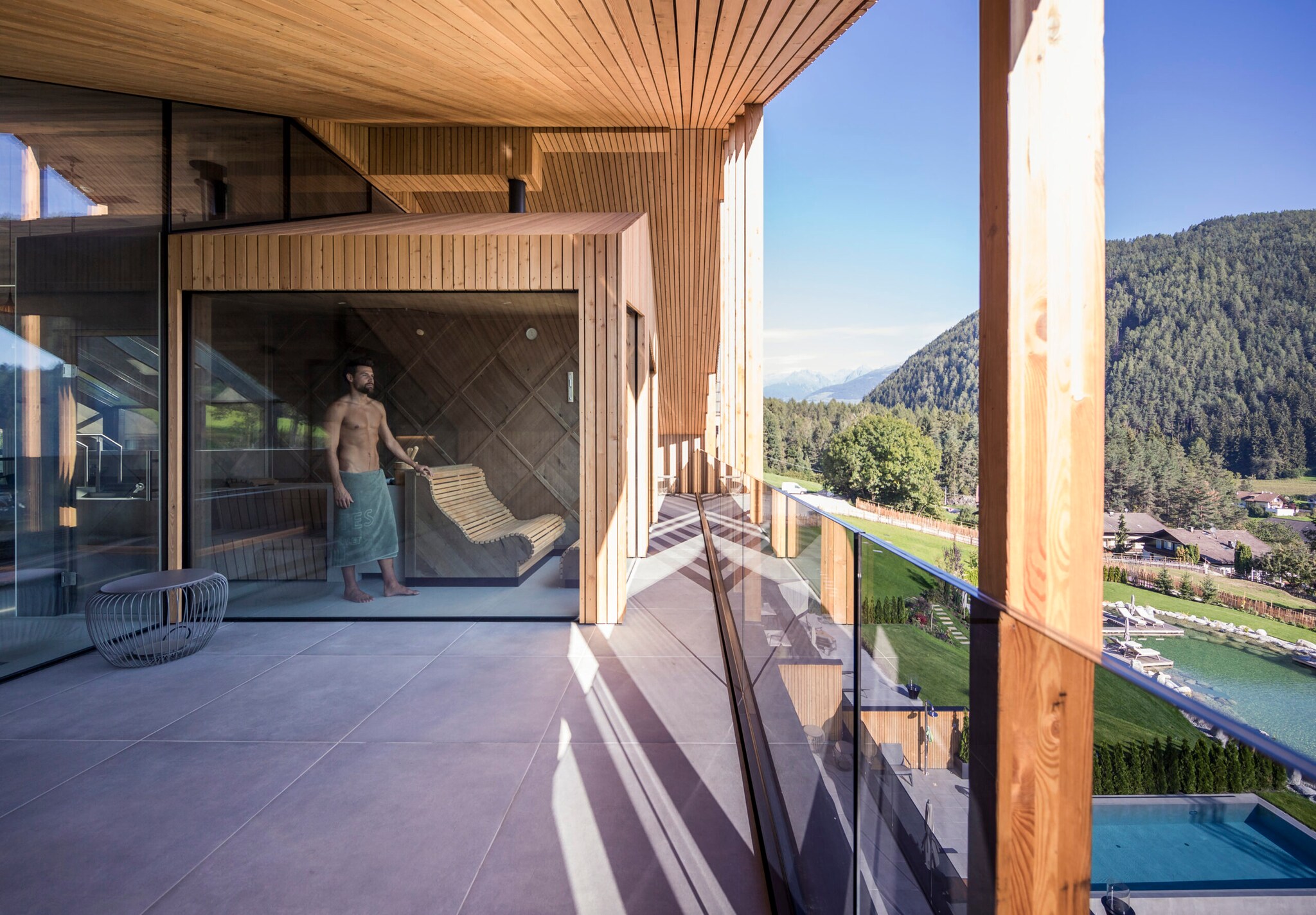 Ein Mann im Badehandtuch steht in einer modernen Holzsauna mit Glasfront auf einer großen Terrasse mit Blick auf eine Berglandschaft Ein Mann im Badehandtuch steht in einer modernen Holzsauna mit Glasfront auf einer großen Terrasse mit Blick auf eine Berglandschaft