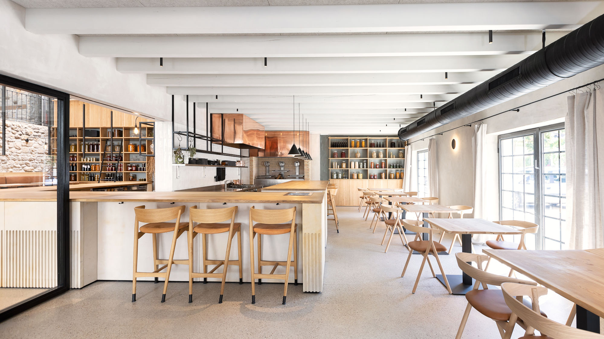 Helles Restaurant mit Holzstühlen und Tischen, offener Decke mit sichtbaren Balken, großer Fensterfront und Regalen mit Büchern im Hintergrund.