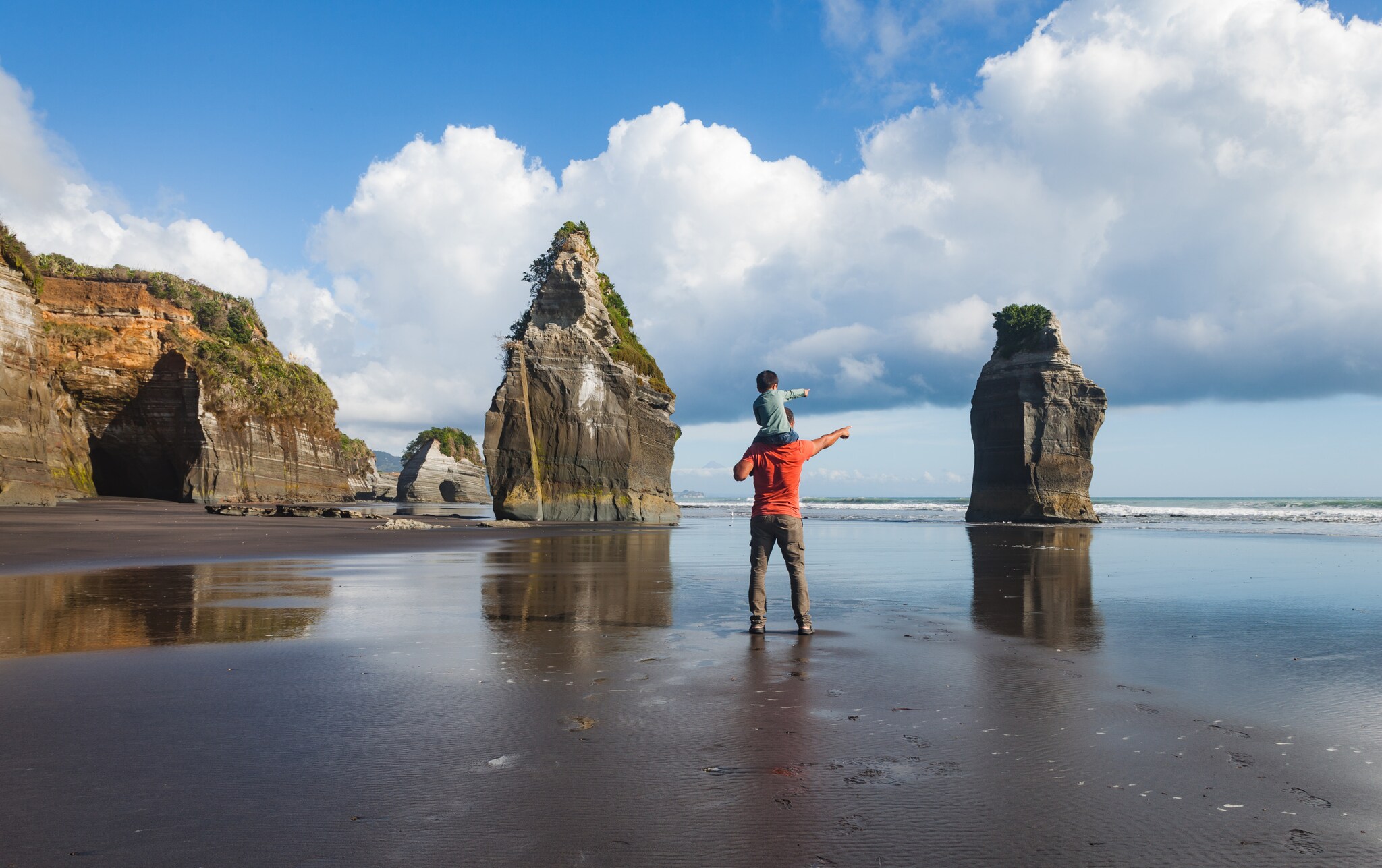 Vater trägt seinen kleinen Sohn auf der Schulter, beide bestaunen die felsige Küstenlandschaft des Three Sister Beach in North Taranaki.