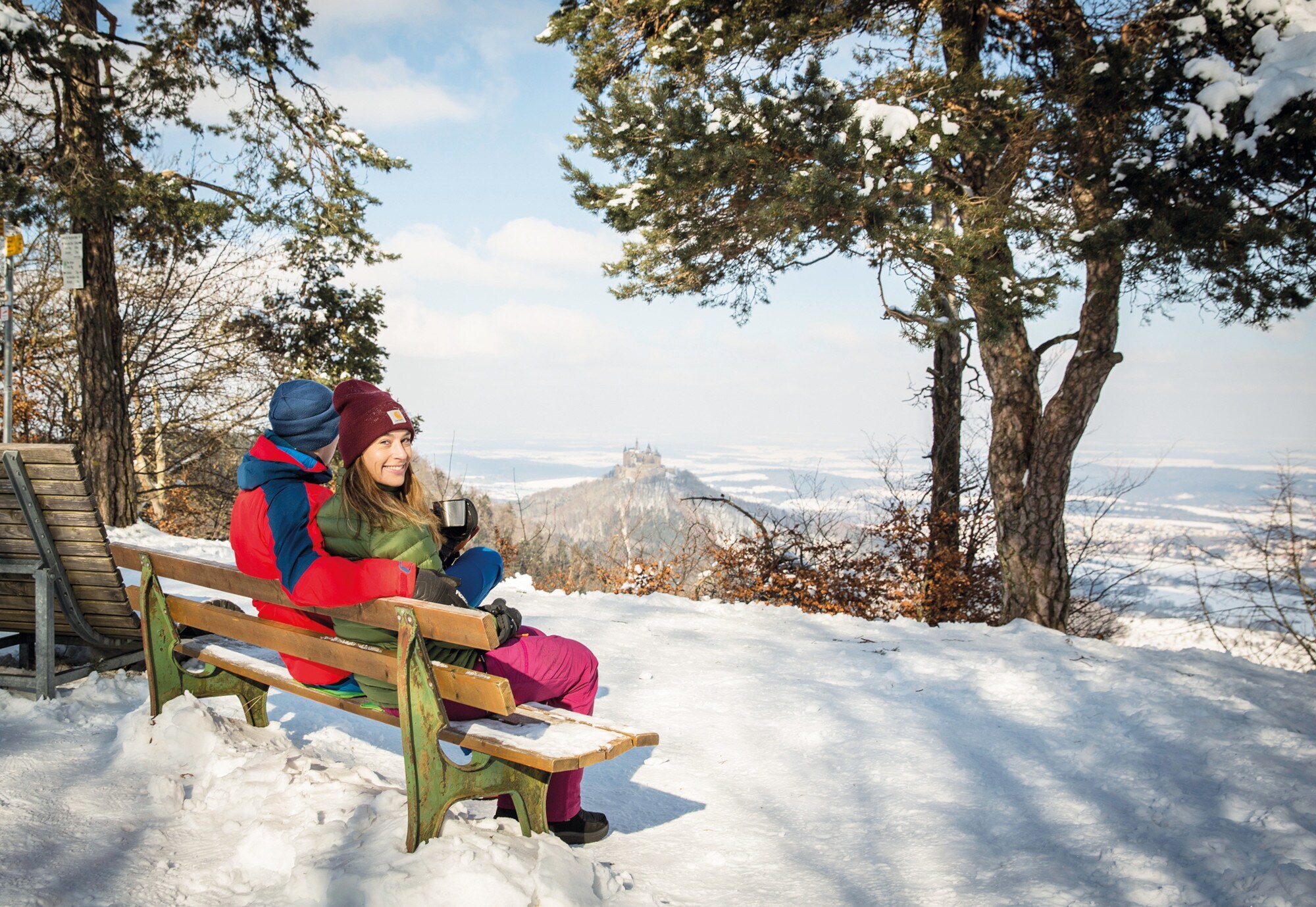 Ein Paar sitzt auf einer verschneiten Bank und genießt den Blick in die umliegende weiße Winterlandschaft mit einem Thermobecher in der Hand