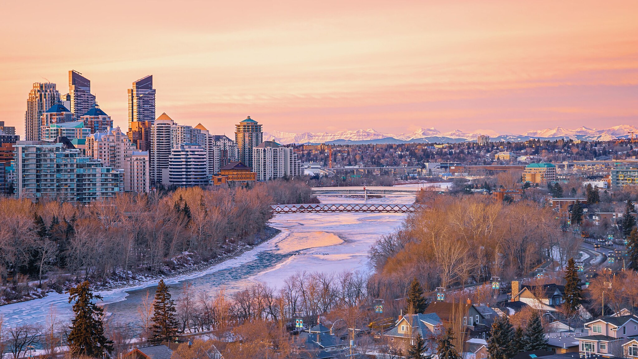 Stadtpanorama von Calgary mit Skyline am gefrorenen See vor schneebedeckter Bergkette am Horizont im violettfarbenen Winterlicht bei Sonnenaufgang.