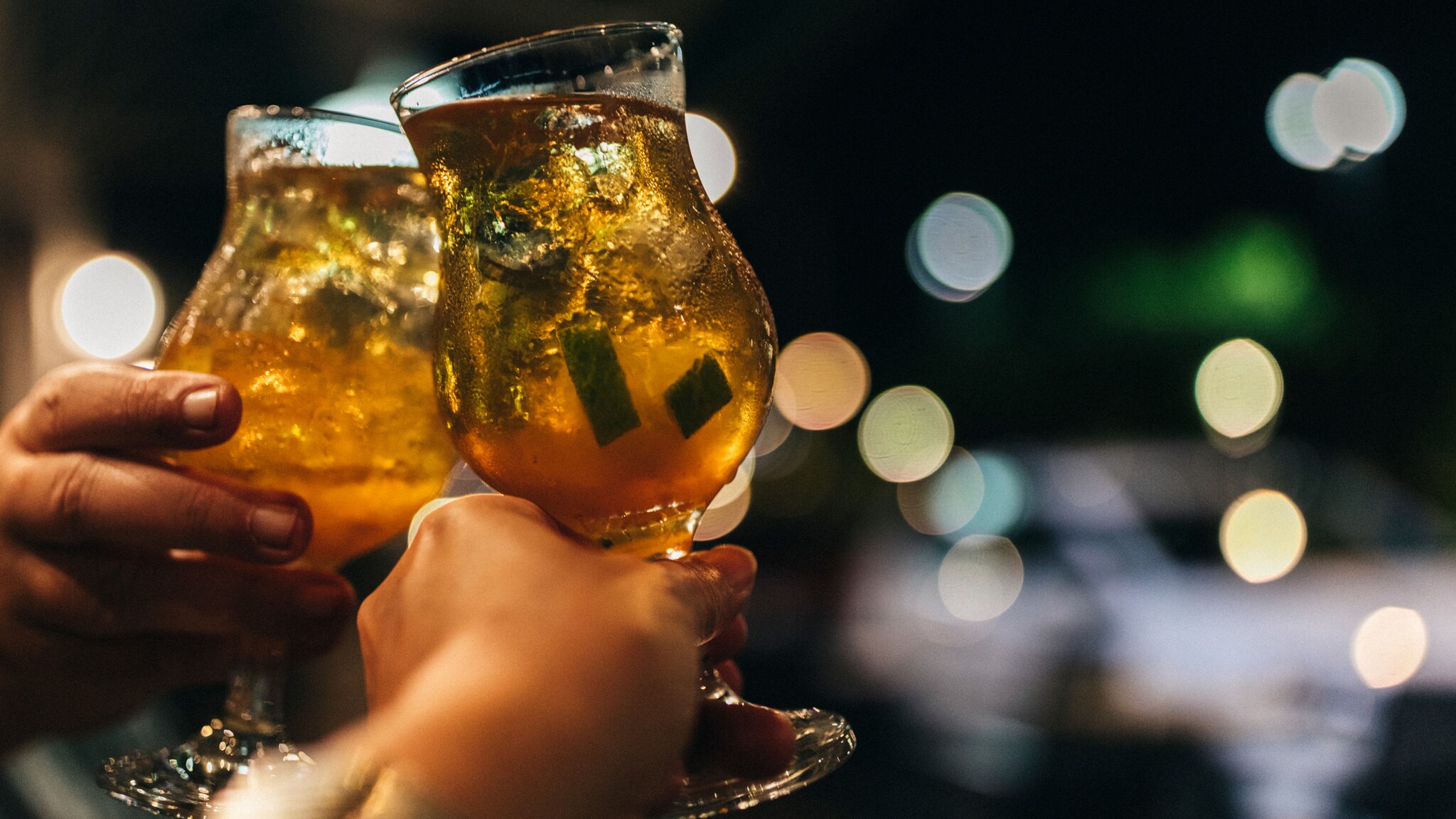 Zwei Hände stoßen mit Gläsern voller Eis und goldfarbenem Getränk bei Nacht an Zwei Hände stoßen mit Gläsern voller Eis und goldfarbenem Getränk bei Nacht an