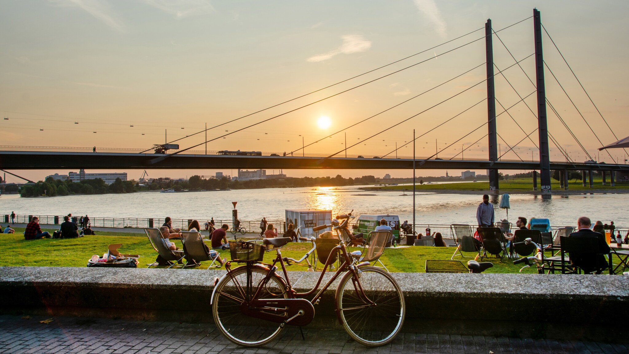Ein rotes Fahrrad steht auf dem Bürgersteig, im Hintergrund ist eine Brücke und der Sonnenuntergang über dem Wasser zu sehen. Menschen entspannen auf der Wiese.