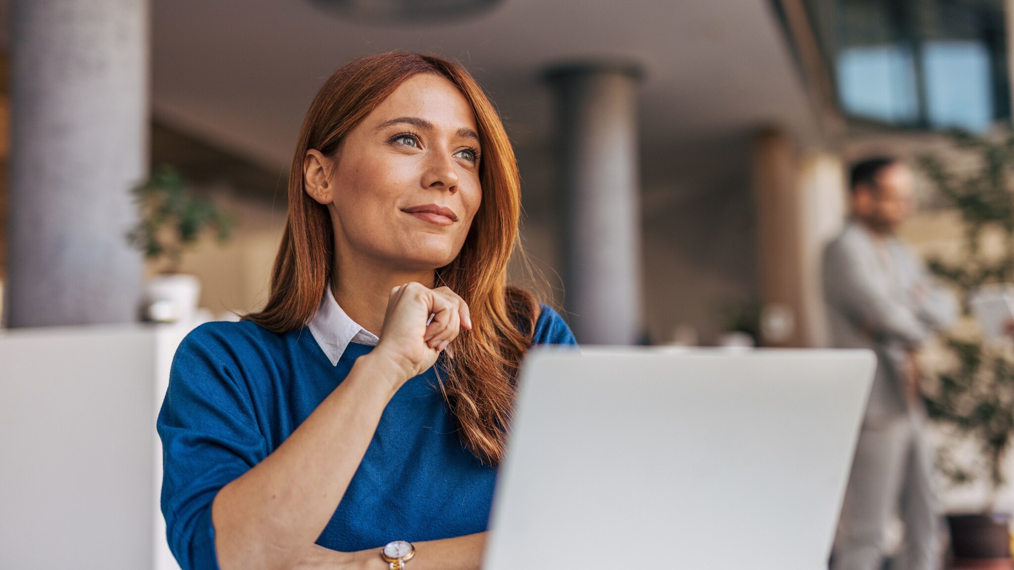 Eine Frau im blauen Pullover sitzt vor einem Laptop und schaut zufrieden lächelnd in ein Büro. Eine Frau im blauen Pullover sitzt vor einem Laptop und schaut zufrieden lächelnd in ein Büro.