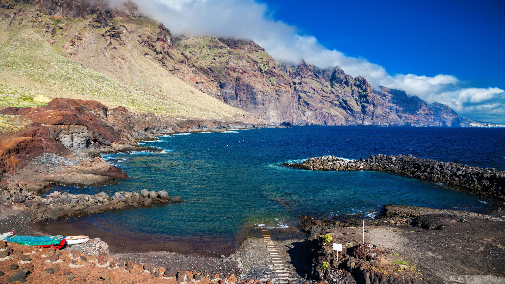 Küstenlandschaft auf Teneriffa mit steilen Felsen, blauem Meer und bewölktem Himmel, im Vordergrund ein kleiner Steg und felsige Uferbereiche.