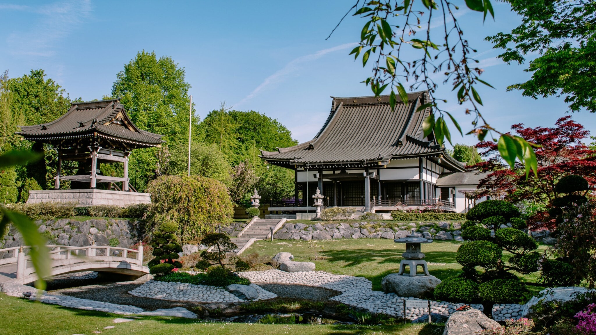 Japanischer Garten mit zwei traditionellen Gebäuden, einem Pavillon und einem Haus mit geschwungenem Dach. Grüne Bäume und gepflegte Sträucher umgeben den Garten. Japanischer Garten mit zwei traditionellen Gebäuden, einem Pavillon und einem Haus mit geschwungenem Dach. Grüne Bäume und gepflegte Sträucher umgeben den Garten.
