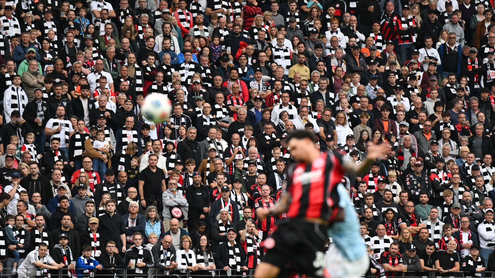 Zuschauende mit schwarz-weiß gestreiften Schals auf einer Tribüne in einem Stadion, im Vordergrund ein Fußballspieler in Unschärfe in einem schwarz-weiß gestreiften Trikot.