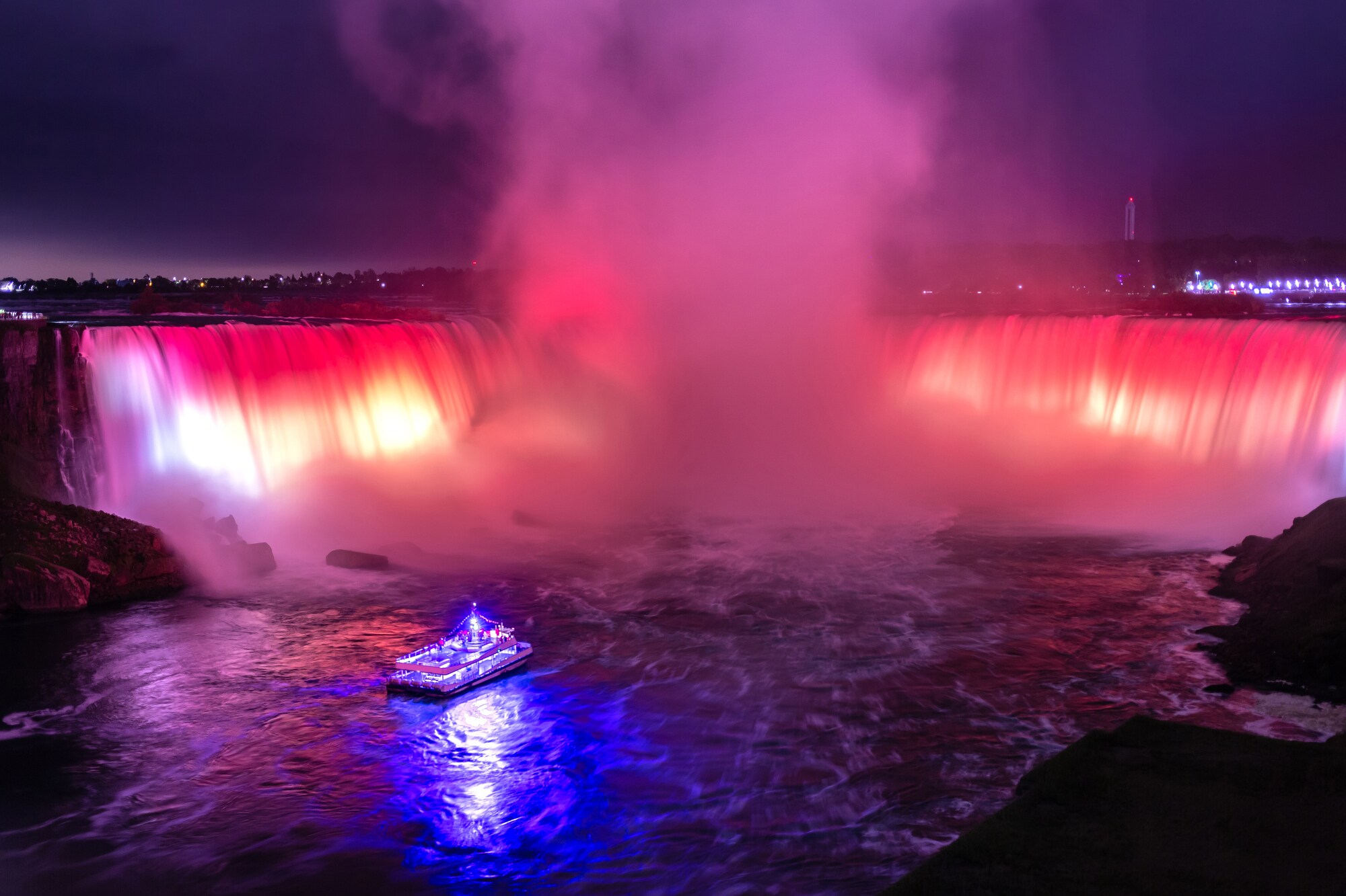 Ein blau beleuchtetes Ausflugsboot vor den rot beleuchteten Niagarafällen bei Nacht.