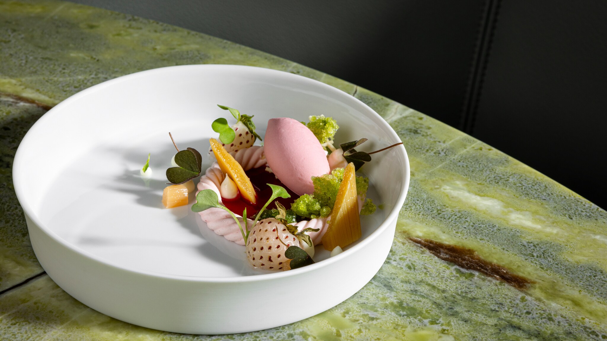 Weißer Teller mit rosa Sorbet, Babymais, weißen Erdbeeren und grünen Kräutern auf grün marmoriertem Tisch.