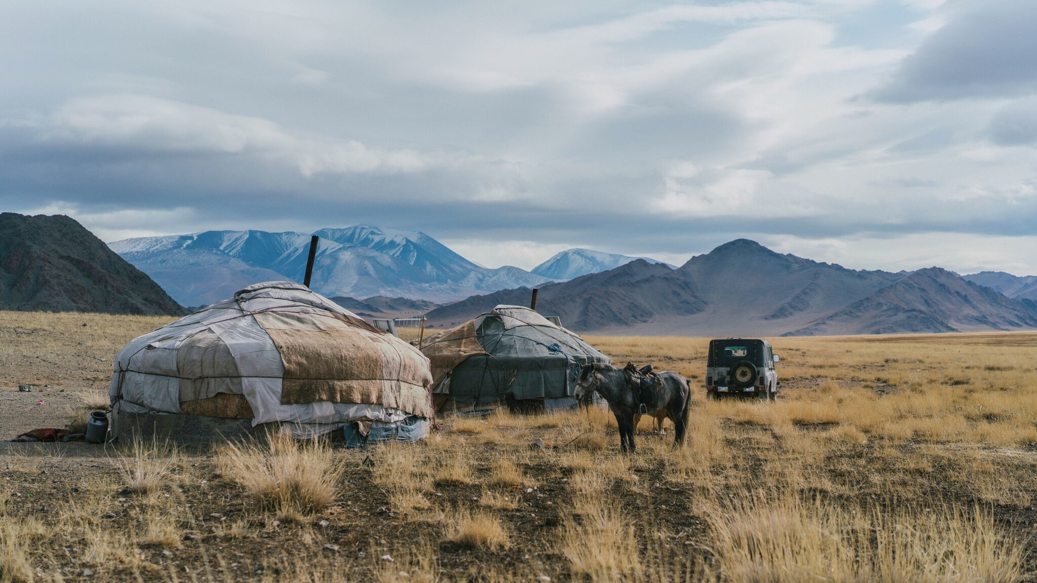 Zwei traditionelle Jurten in der Wüste Gobi vor Gebirgspanorama, daneben steht ein Esel und ein Jeep parkt in der Ferne.