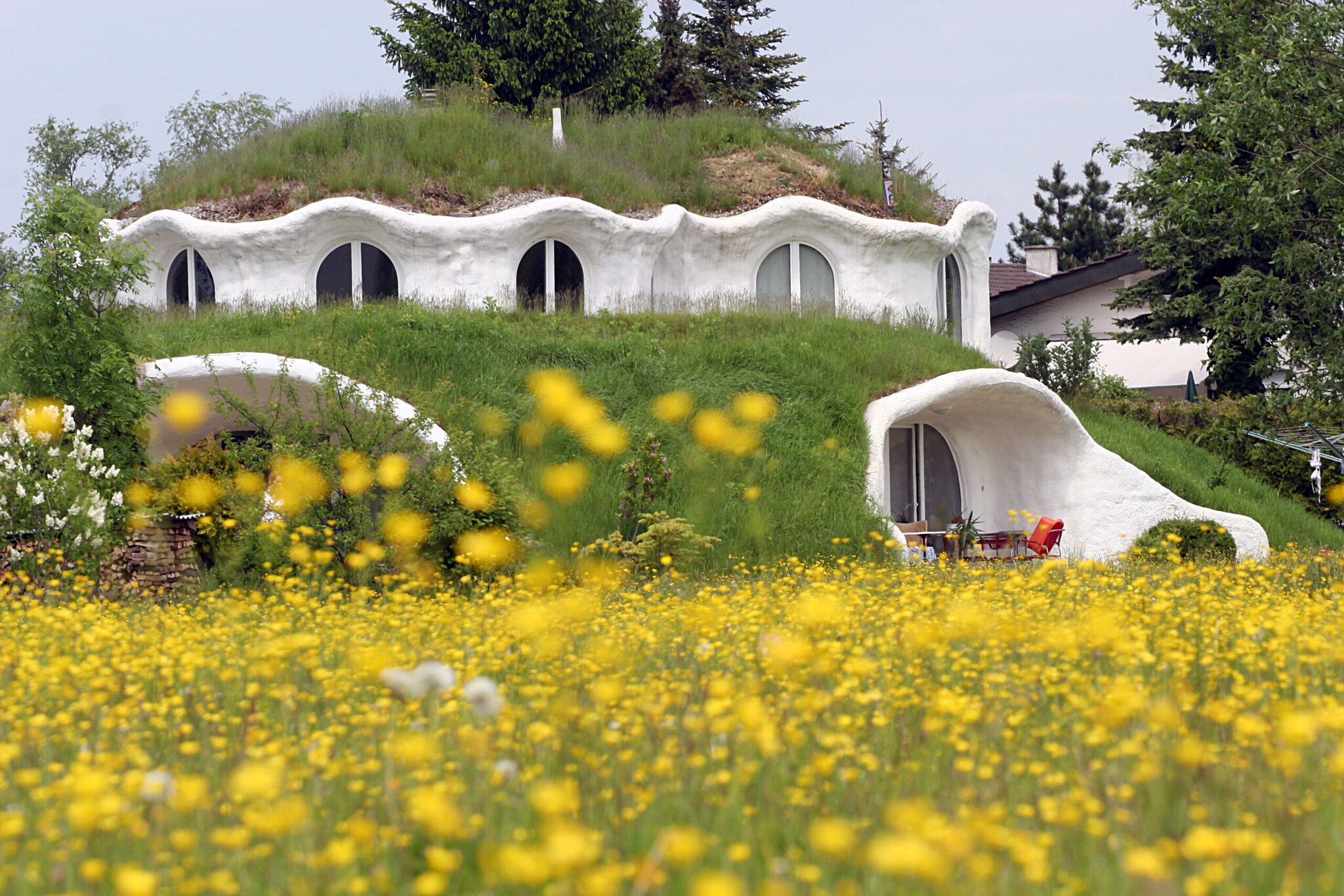 Großes, zweistöckiges Erdhügelhaus mit weißen, organisch-geformten Wänden und Grasdächern, im Vordergrund eine Blumenwiese