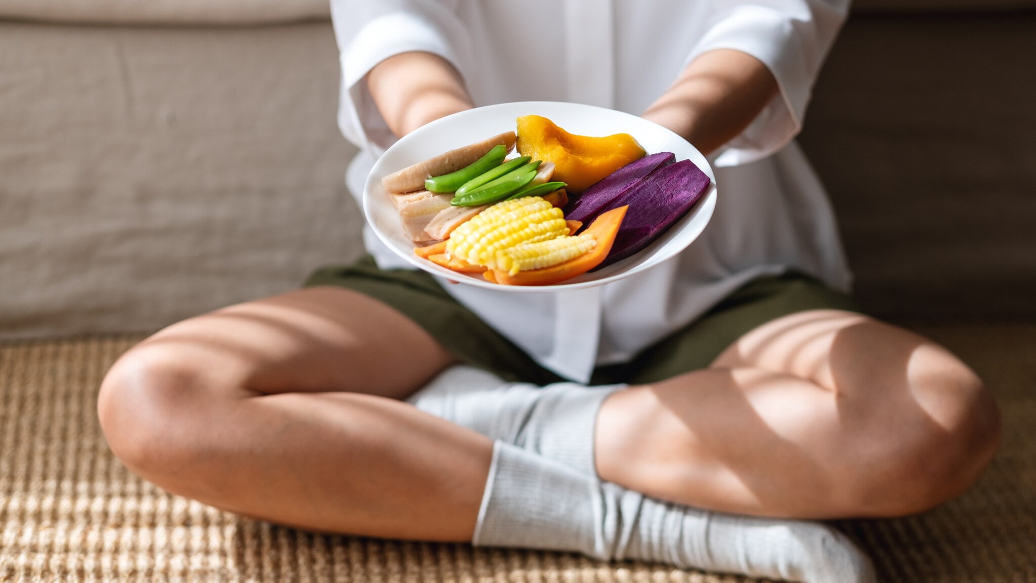 Person sitzt im Schneidersitz und hält einen Teller mit buntem, geschnittenem Gemüse, darunter Mais, Süßkartoffel und grüne Bohnen. Person sitzt im Schneidersitz und hält einen Teller mit buntem, geschnittenem Gemüse, darunter Mais, Süßkartoffel und grüne Bohnen.