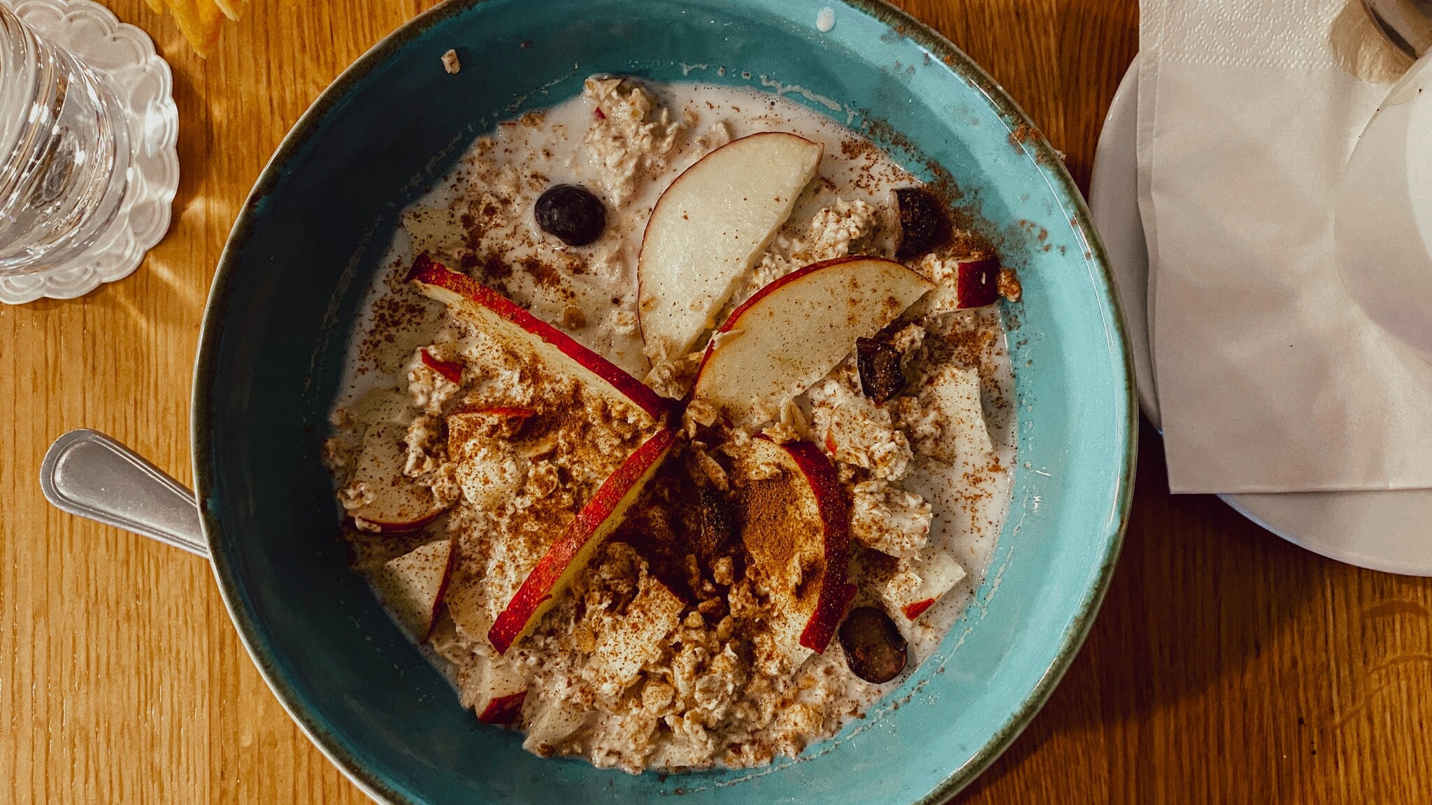 Blaue Schüssel mit Porridge, garniert mit Apfelscheiben, Blaubeeren und Zimt, auf Holztisch mit Glas Wasser und Serviette. Blaue Schüssel mit Porridge, garniert mit Apfelscheiben, Blaubeeren und Zimt, auf Holztisch mit Glas Wasser und Serviette.