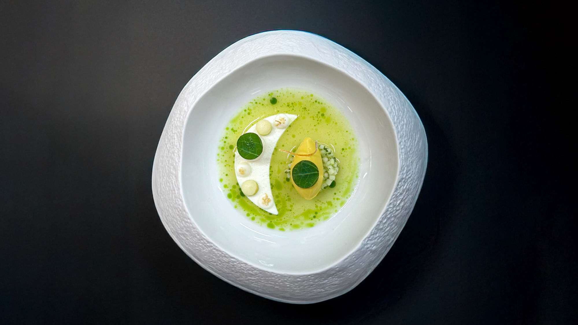 Weißer Teller mit strukturierter Außenkante, darauf halbmondförmige weiße Creme, gelbe Kugel und grüne Soße mit kleinen grünen Blättern. Weißer Teller mit strukturierter Außenkante, darauf halbmondförmige weiße Creme, gelbe Kugel und grüne Soße mit kleinen grünen Blättern.