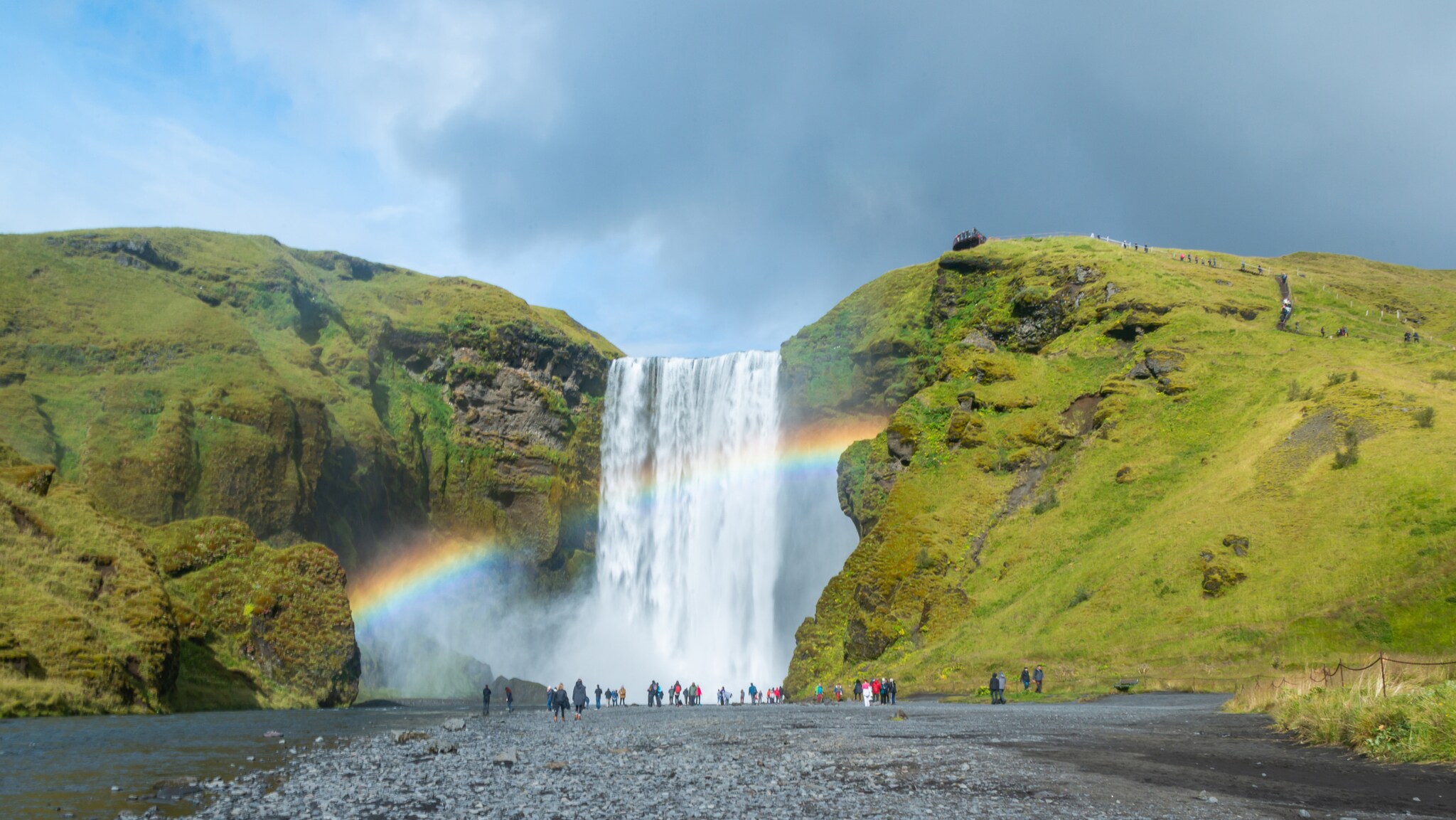 Wasserfall zwischen grünen Felsen mit Regenbogen und Menschen am Ufer unter bewölktem Himmel. Wasserfall zwischen grünen Felsen mit Regenbogen und Menschen am Ufer unter bewölktem Himmel.