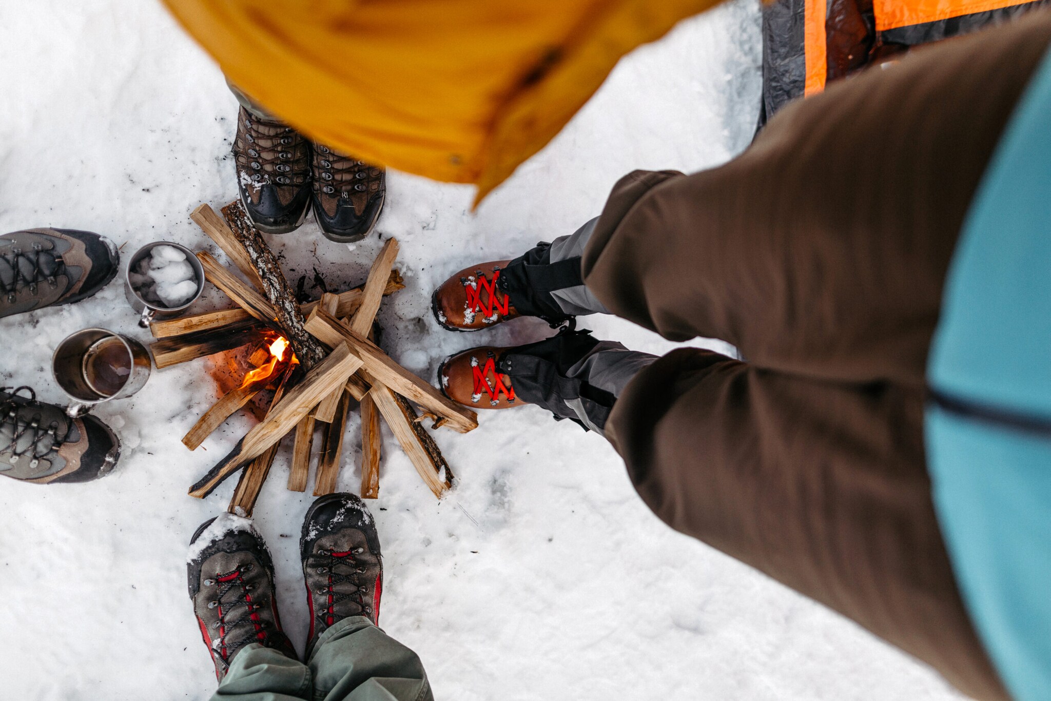 Die Schuhe von vier Personen, die im Schnee um ein Feuer stehen, von oben fotografiert