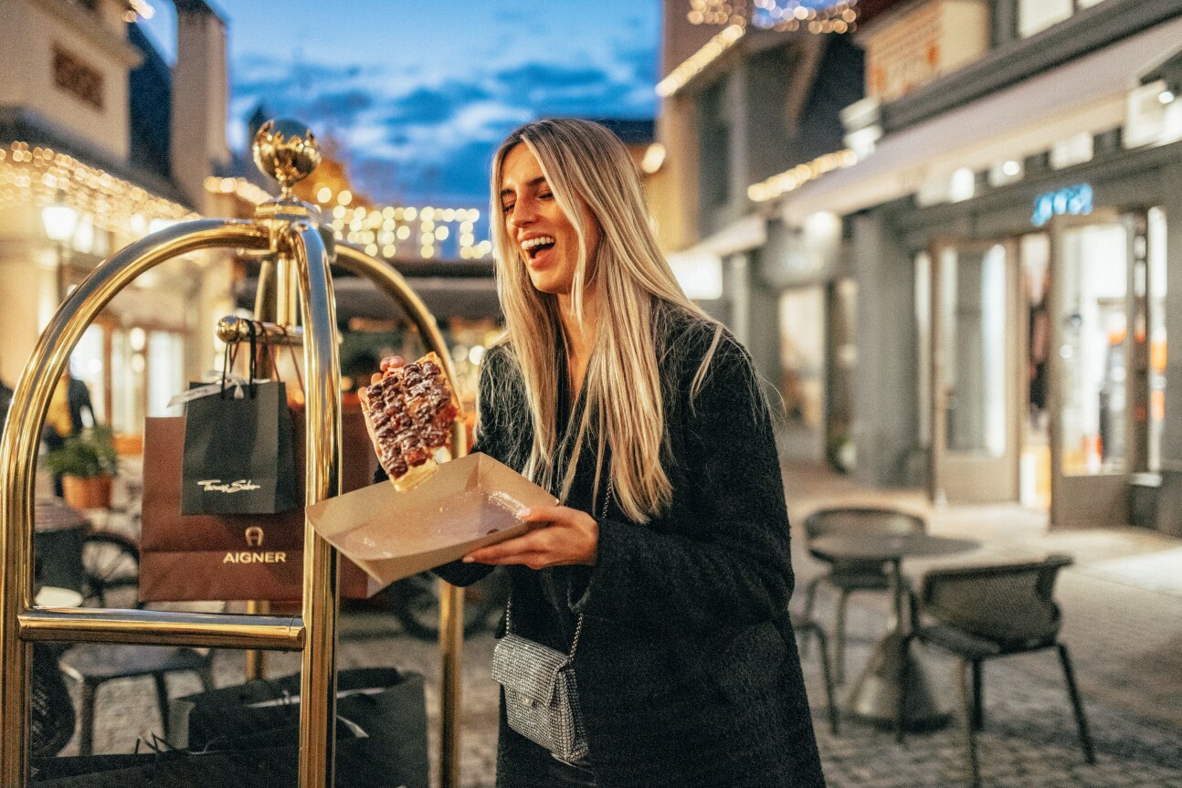 Eine lachende Frau mit einer frischen Waffel in der Hand, neben ihr steht ein Gepäckwagen mit Tüten