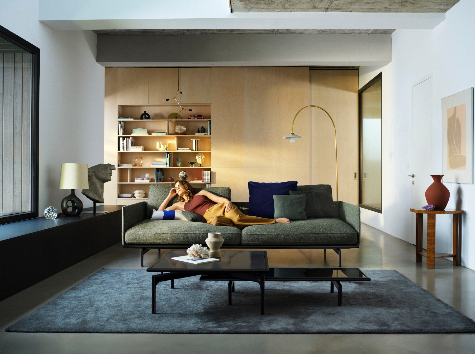 Modern eingerichtetes Wohnzimmer mit Designobjekten, im Zentrum steht ein grünes, geradliniges Sofa, auf dem eine junge Frau liegt Modern eingerichtetes Wohnzimmer mit Designobjekten, im Zentrum steht ein grünes, geradliniges Sofa, auf dem eine junge Frau liegt