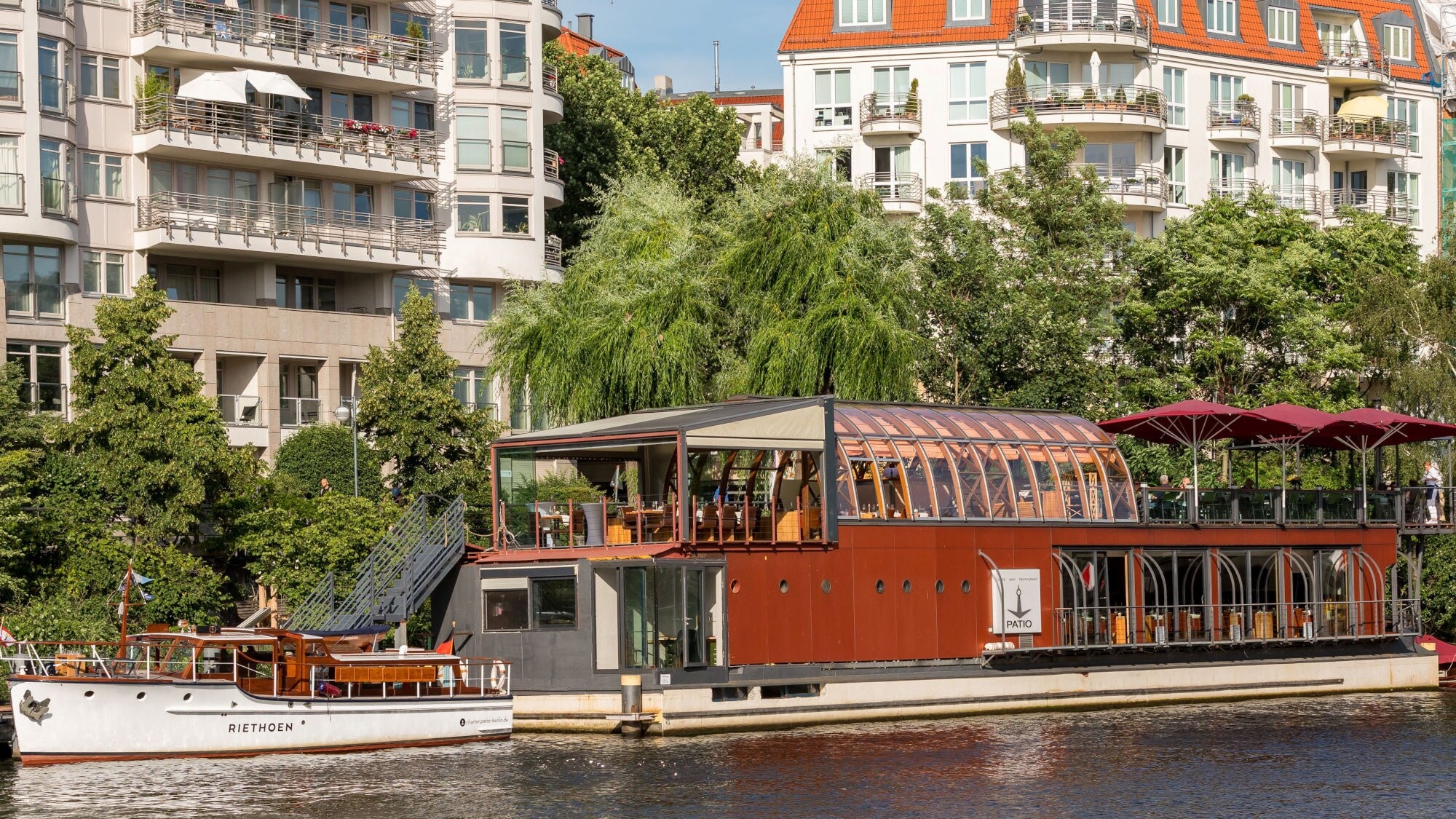 Ein Restaurantboot mit großen Fenstern und einem roten Dach liegt am Ufer eines Flusses. Im Hintergrund sind moderne Wohngebäude und Bäume sichtbar. Ein Restaurantboot mit großen Fenstern und einem roten Dach liegt am Ufer eines Flusses. Im Hintergrund sind moderne Wohngebäude und Bäume sichtbar.