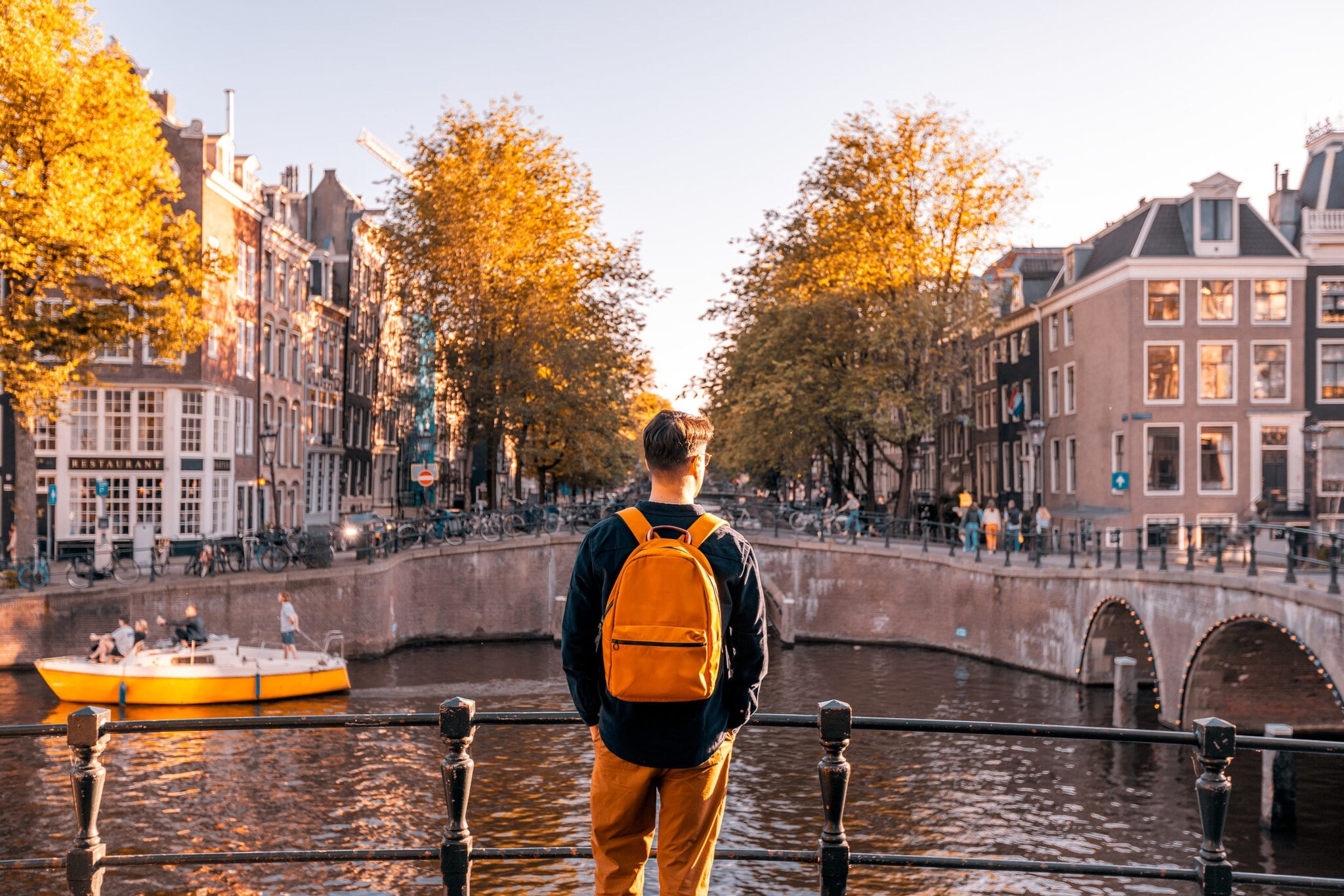 Rückansicht einer Person mit Rucksack, die an einem sonnigen Tag an einem Kanal in Amsterdam steht, auf der anderen Seite Häuser. Rückansicht einer Person mit Rucksack, die an einem sonnigen Tag an einem Kanal in Amsterdam steht, auf der anderen Seite Häuser.