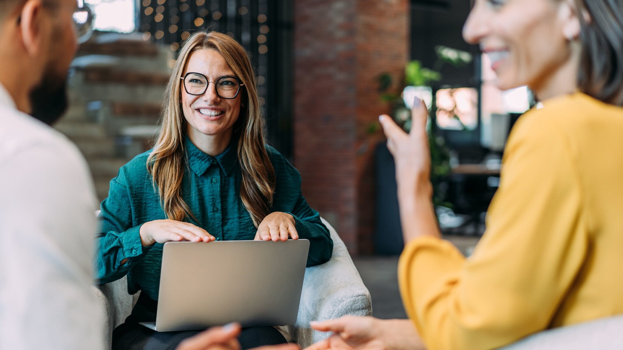 Drei Personen sitzen beieinander und kommunizieren lächelnd, eine Frau hat einen geöffneten Laptop auf ihrem Schoß. Drei Personen sitzen beieinander und kommunizieren lächelnd, eine Frau hat einen geöffneten Laptop auf ihrem Schoß.
