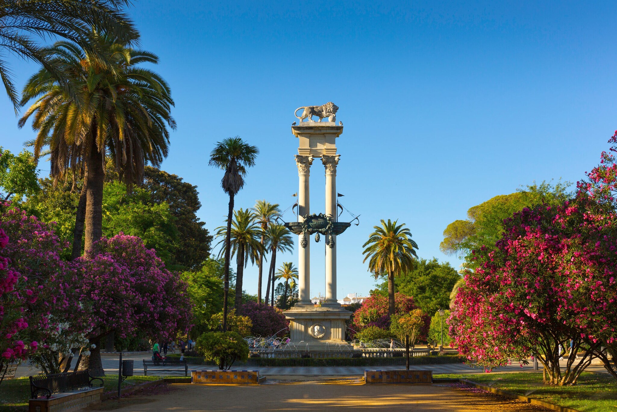 Blick auf das hohe Kolumbus-Denkmal, umgeben von grünen Bäumen, Palmen und pink blühenden Büschen.