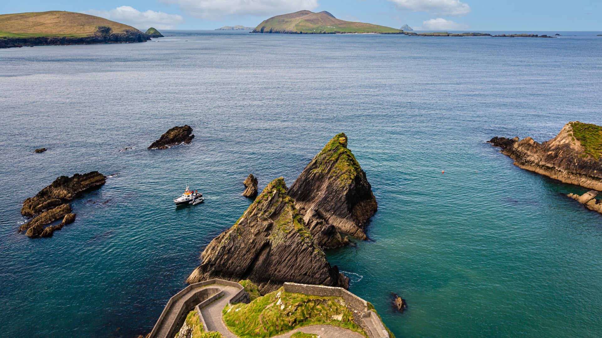 Blick von oben auf eine Küstenlandschaft mit einem geschwungenen Steinweg, Felsen im Wasser und Inseln im Hintergrund.