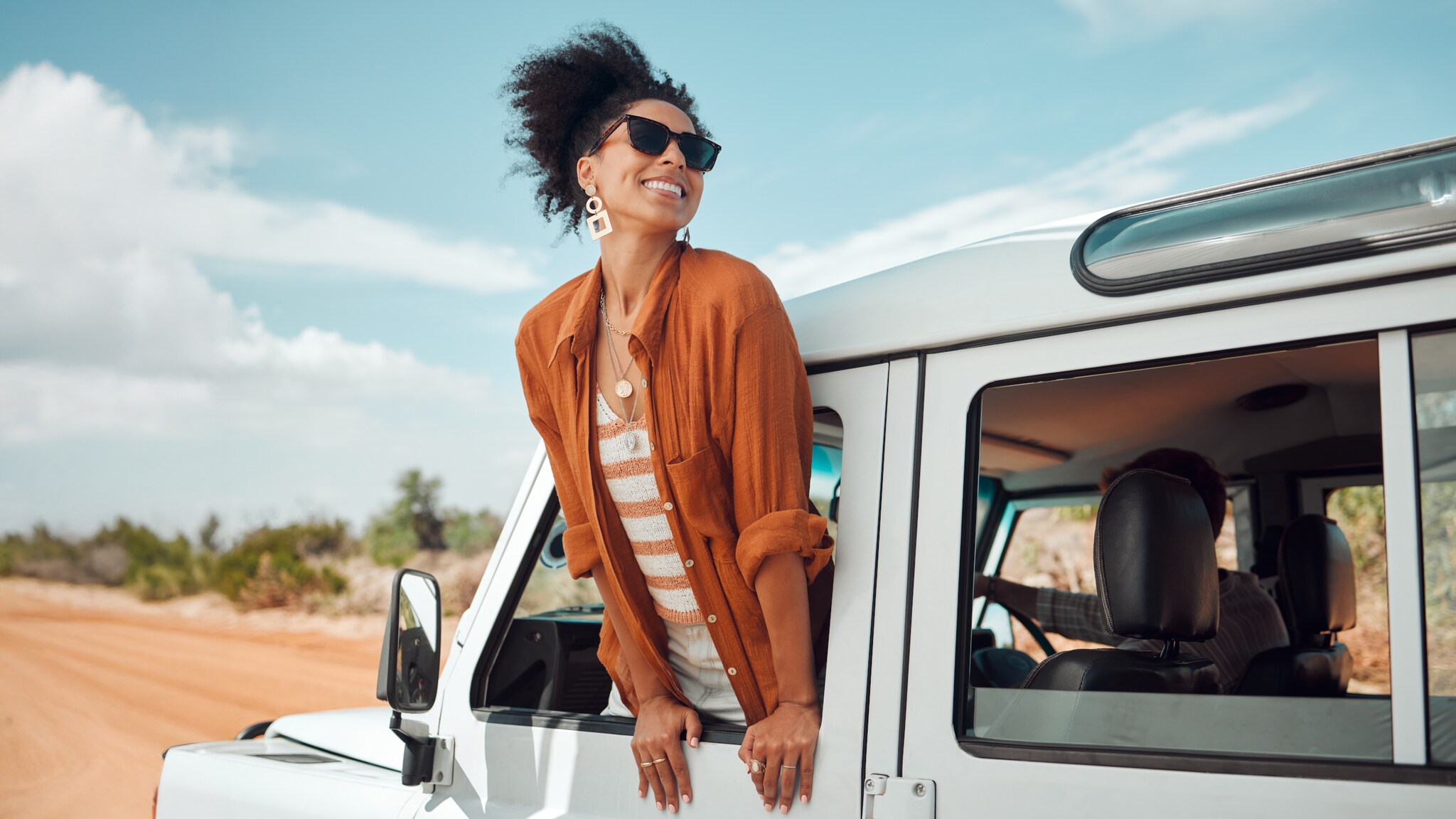 Eine Frau mit lockigem Haar lehnt aus dem Fenster eines weißen Geländewagens. Sie trägt eine orangefarbene Bluse und eine gestreifte Bluse darunter. Im Hintergrund ist eine Landschaft mit Bäumen und einem blauen Himmel zu sehen.