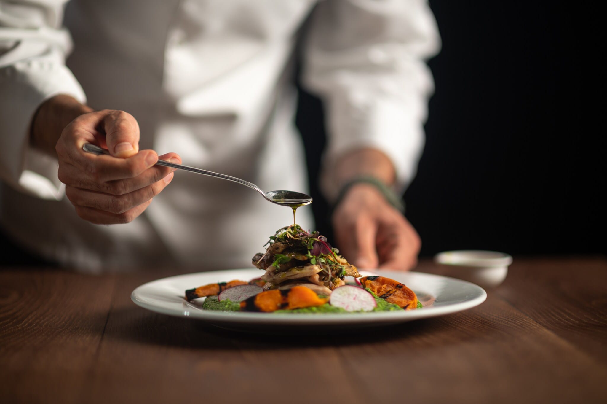 Die Hände eines Kochs, der Soße aus dem Löffel auf eine Mahlzeit auf einem weißen Teller gießt. Die Hände eines Kochs, der Soße aus dem Löffel auf eine Mahlzeit auf einem weißen Teller gießt.