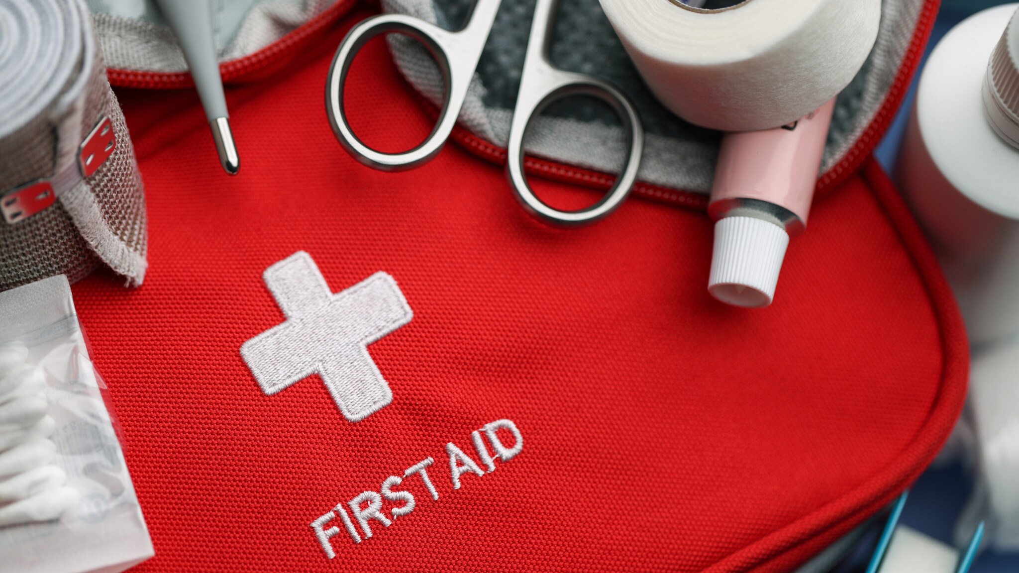 Roter Erste-Hilfe-Kasten mit einem weißen Kreuz und dem Text 'FIRST AID'. Umgeben von Scheren, Binden, Watte und verschiedenen medizinischen Utensilien.