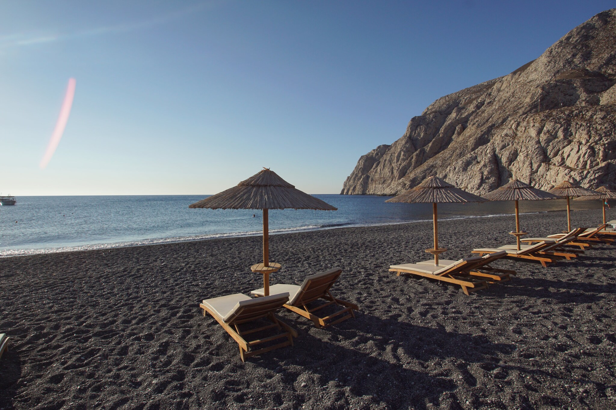 Liegen und Sonnenschirme an einem menschenleeren Strand mit schwarzem Sand in einer Bucht.