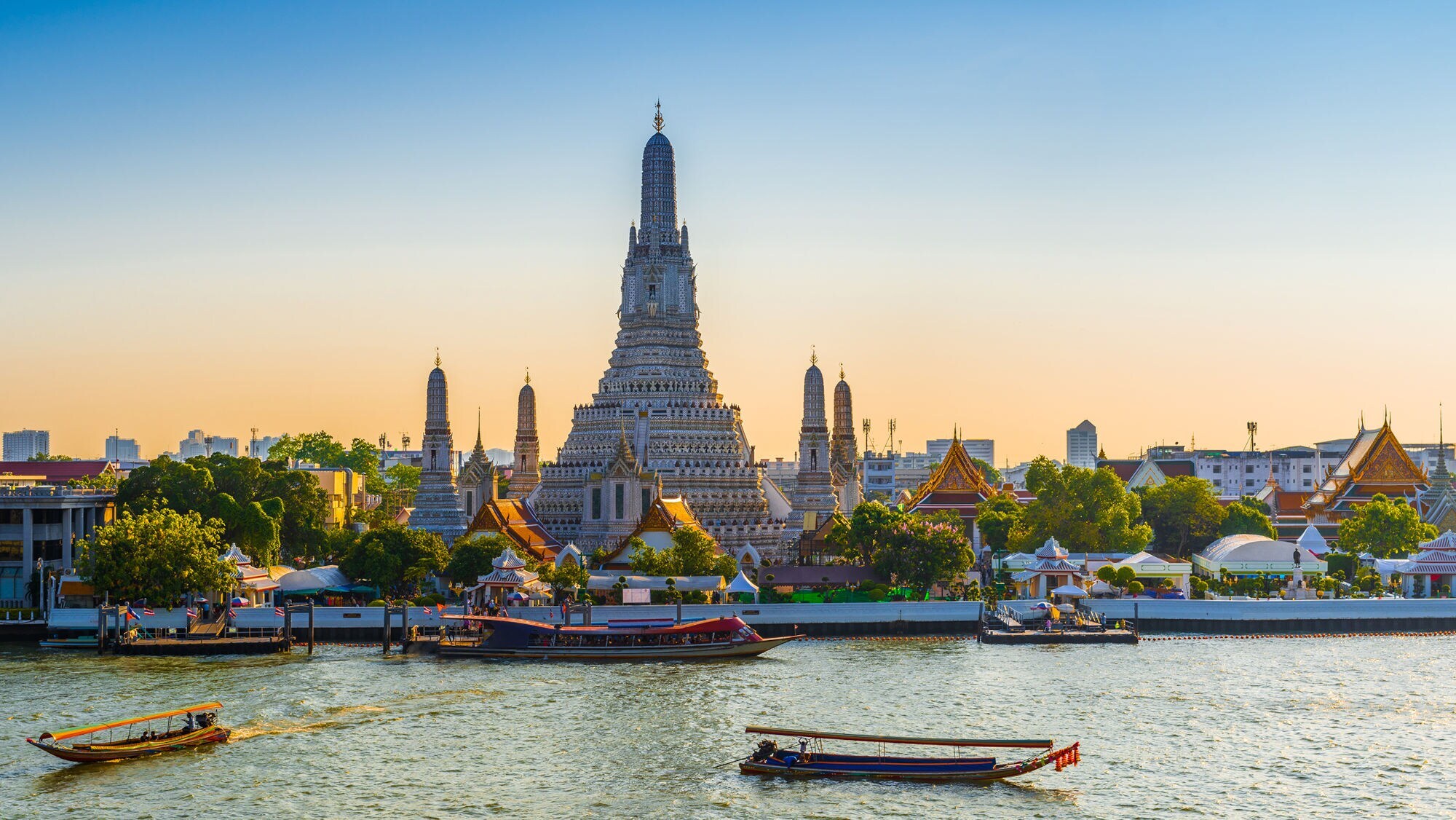 Vor einem großen Fluss mit Booten ragt ein großer Tempel empor, er ist umgeben von kleineren Türmen und Häusern mit spitzen Dächern.