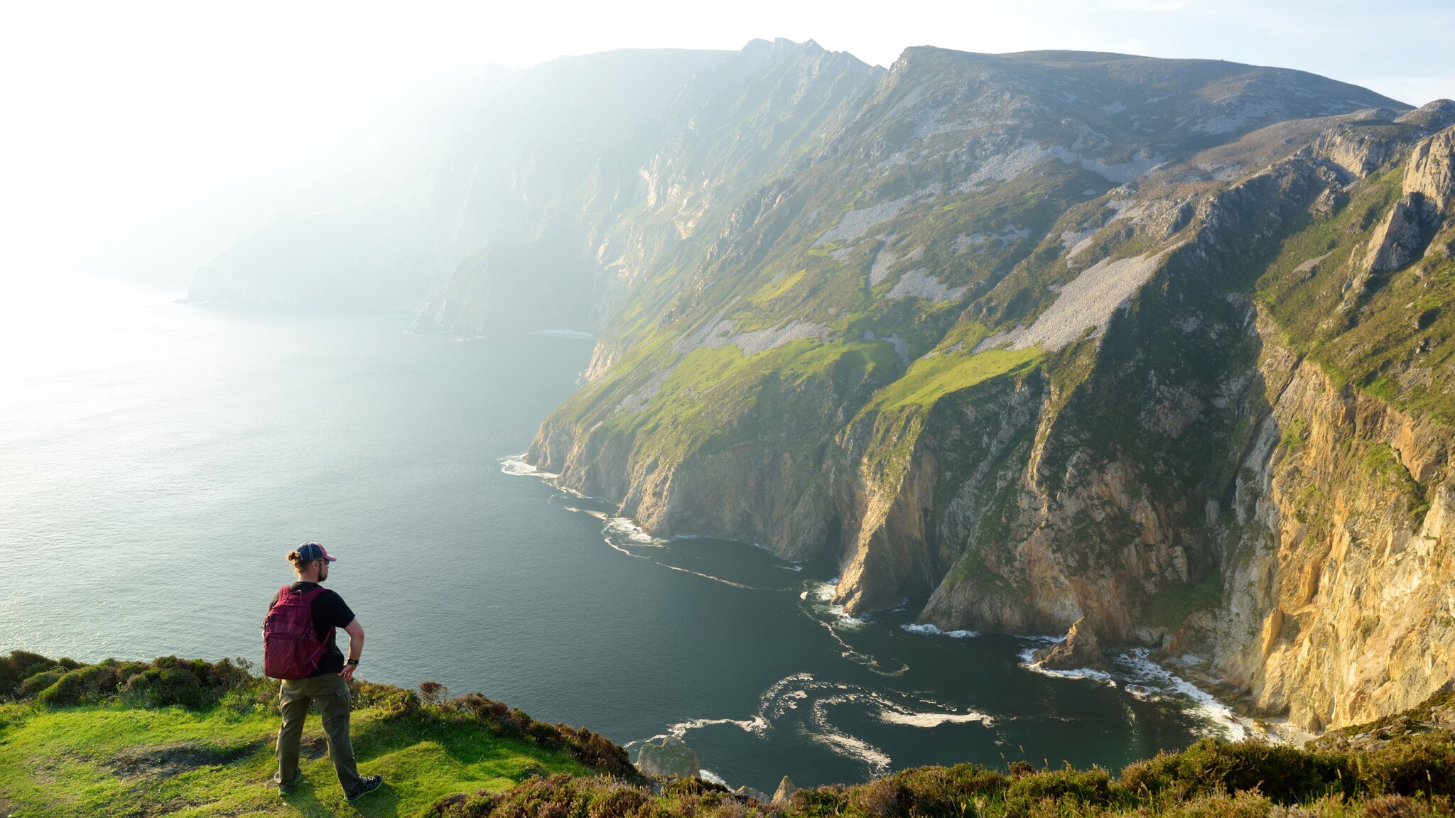 Ein Wanderer mit einem Rucksack steht auf einer grünen Wiese und blickt auf eine steile Küste mit Klippen und dem Meer im Hintergrund. Ein Wanderer mit einem Rucksack steht auf einer grünen Wiese und blickt auf eine steile Küste mit Klippen und dem Meer im Hintergrund.