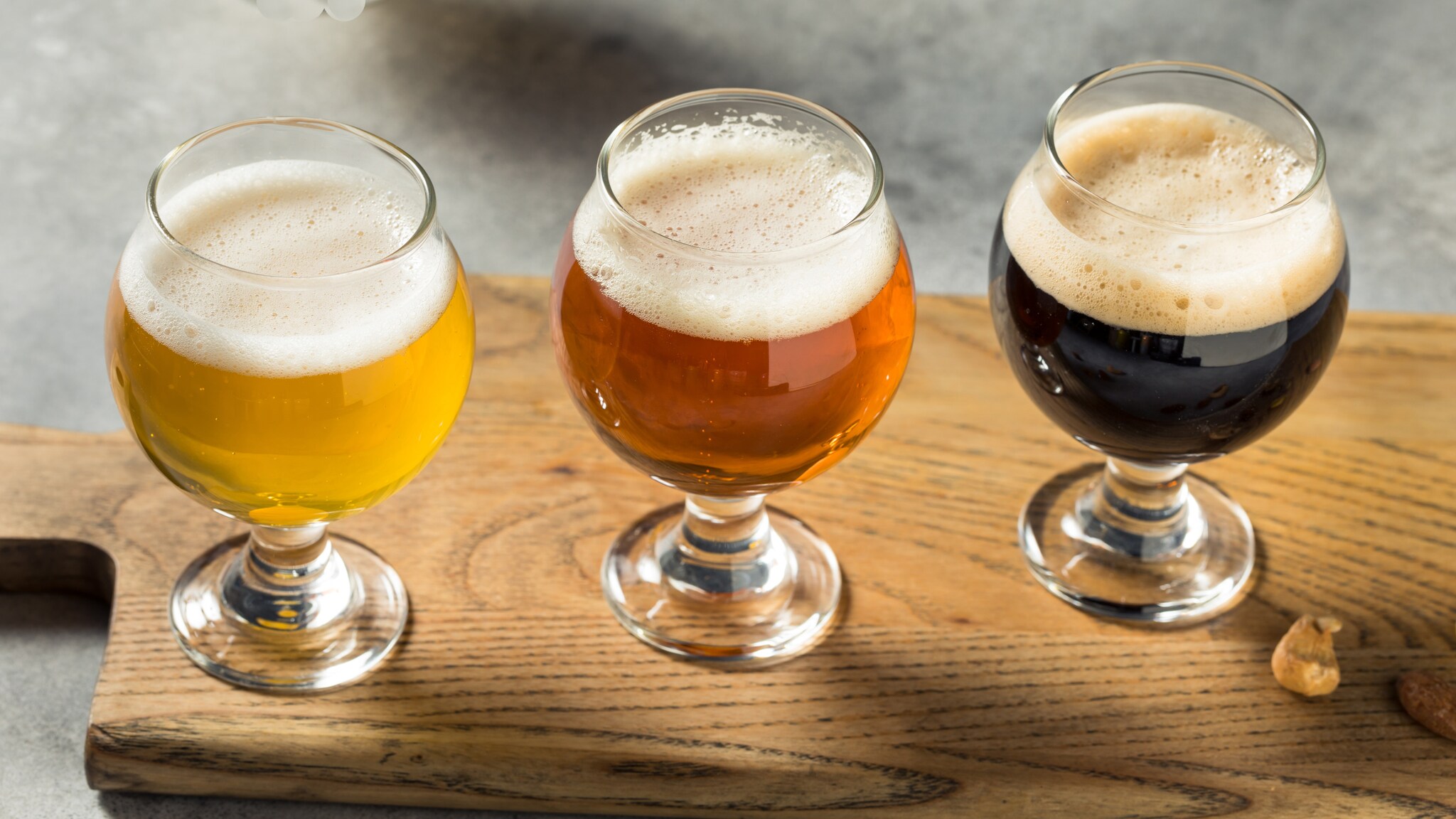Drei Gläser mit verschiedenen Biersorten auf einem Holzbrett. Links ein helles Bier, in der Mitte ein bernsteinfarbenes Bier und rechts ein dunkles Bier. Schaumkrone auf jedem Glas.