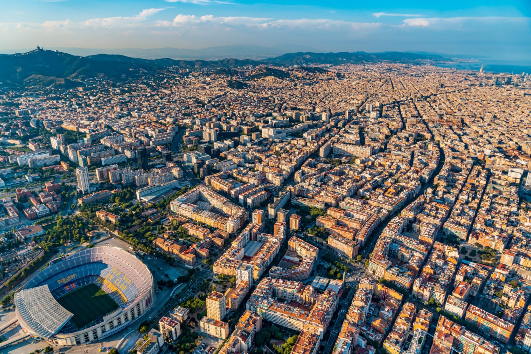 Stadtpanorama von Barcelona mit Fußballstadion aus der Luftperspektive, im Hintergrund eine Hügelkette und Meer.