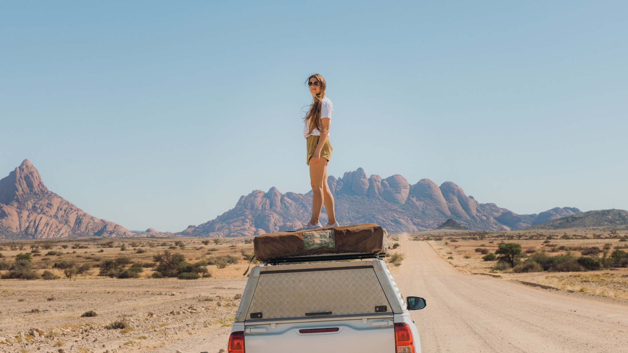 Eine Frau mit Sonnenbrille steht auf einem Auto in Namibia, hinter ihr die Berge der Spitzkoppe.