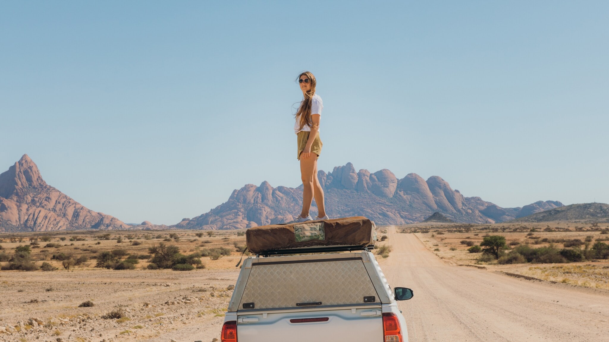 Eine Frau mit Sonnenbrille steht auf einem Auto in Namibia, hinter ihr die Berge der Spitzkoppe. Eine Frau mit Sonnenbrille steht auf einem Auto in Namibia, hinter ihr die Berge der Spitzkoppe.