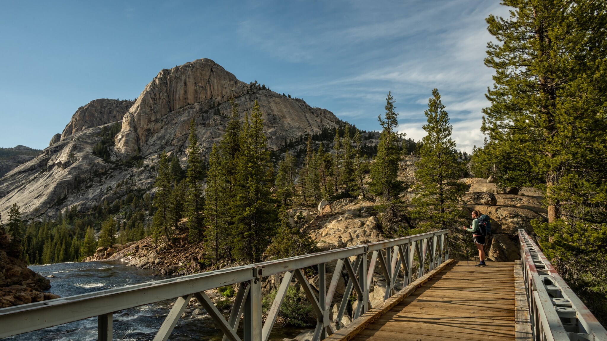 Wanderer mit Rucksack auf einer Holzbrücke über einen Fluss, umgeben von Kiefern und Bergen bei bewölktem Himmel Wanderer mit Rucksack auf einer Holzbrücke über einen Fluss, umgeben von Kiefern und Bergen bei bewölktem Himmel