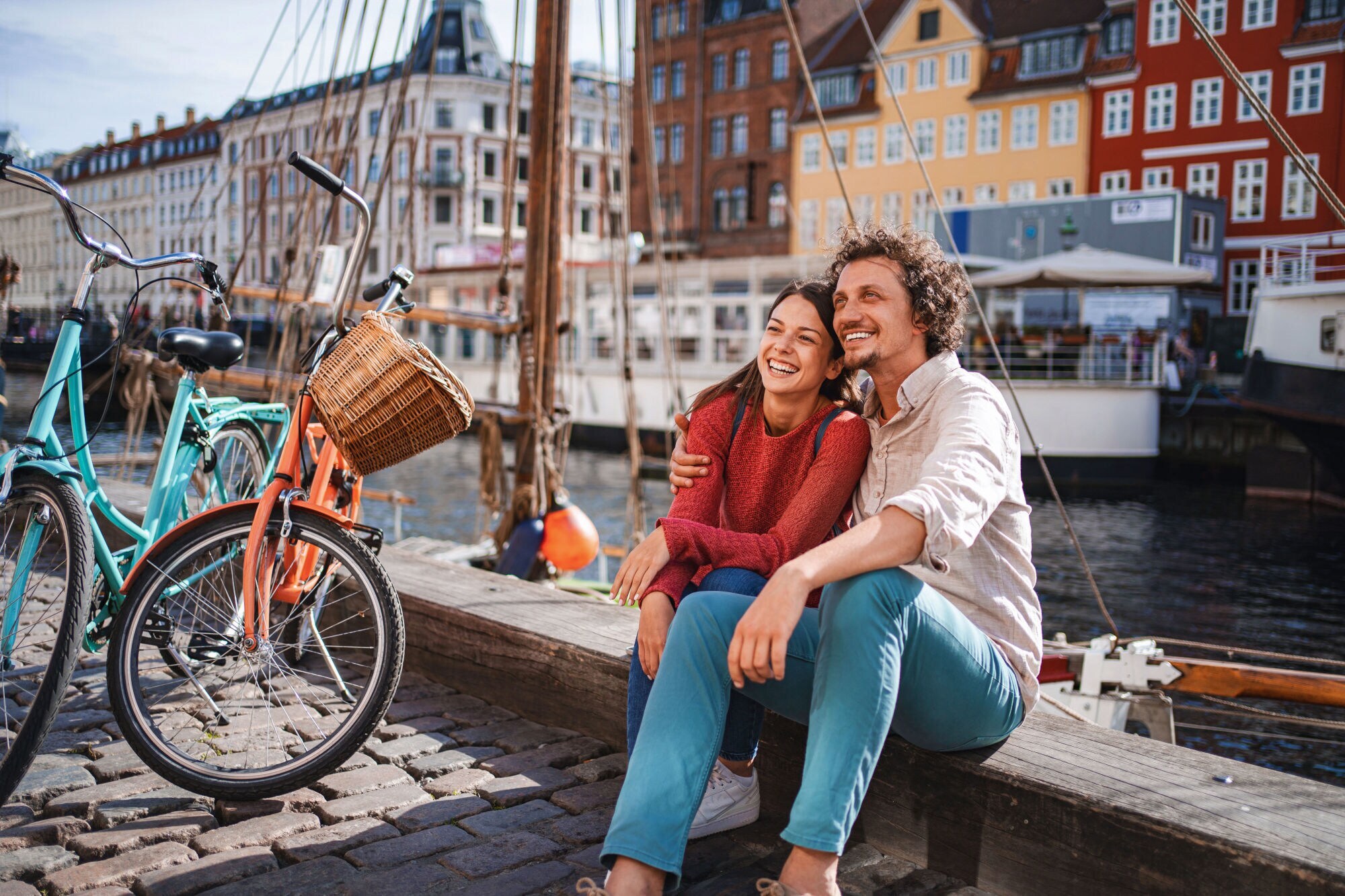 Ein lachendes Paar sitzt eng beieinander auf einer Kaimauer an einem Kanal mit bunten Häusern im Stadtzentrum bei Sonnenschein, neben ihnen stehen zwei Fahrräder auf Kopfsteinpflaster.