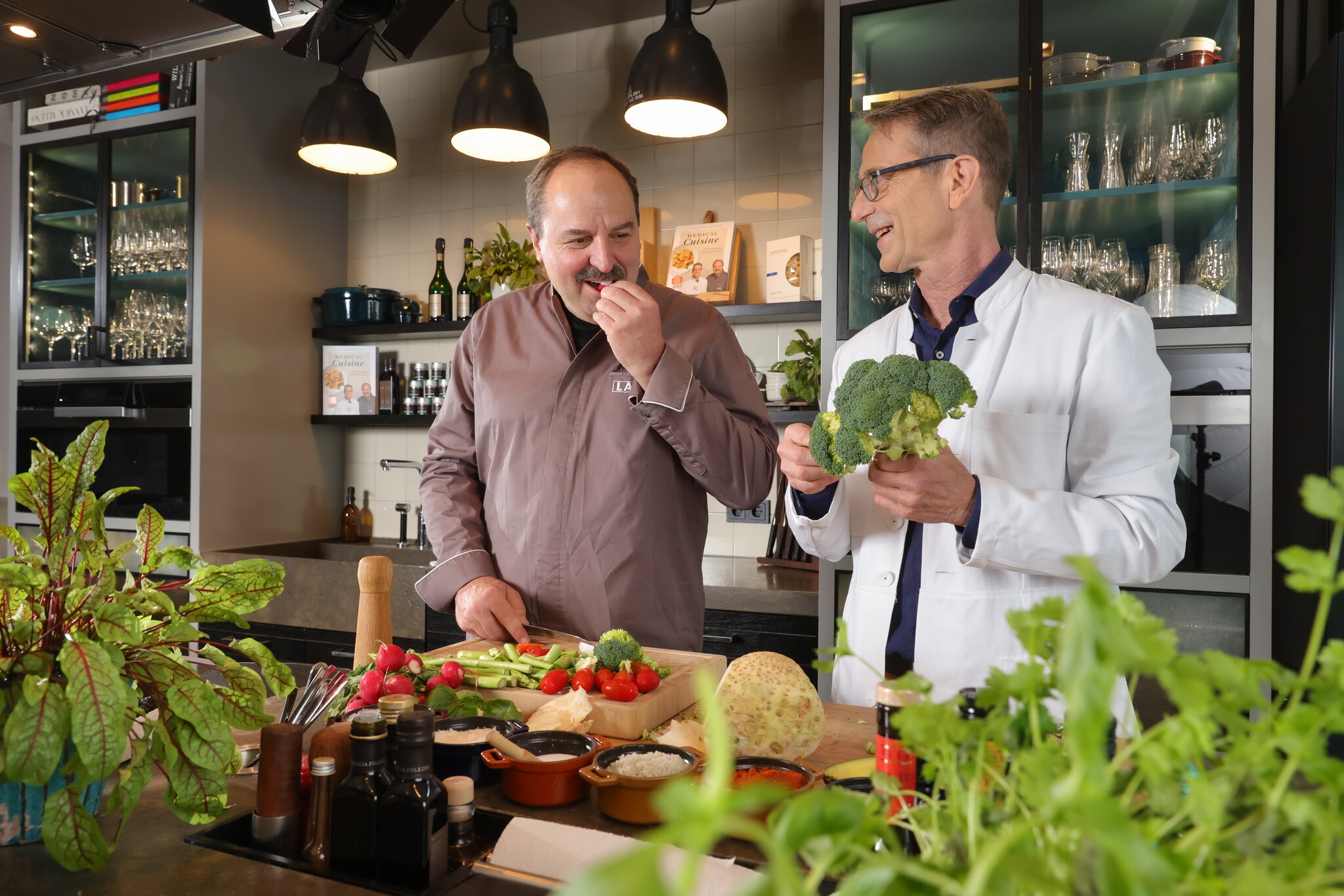 Starkoch Johann Lafer und Ernährungsmediziner Dr. Matthias Riedl stehen nebeneinander in einer Küche vor einer Theke mit rohem Gemüse Starkoch Johann Lafer und Ernährungsmediziner Dr. Matthias Riedl stehen nebeneinander in einer Küche vor einer Theke mit rohem Gemüse