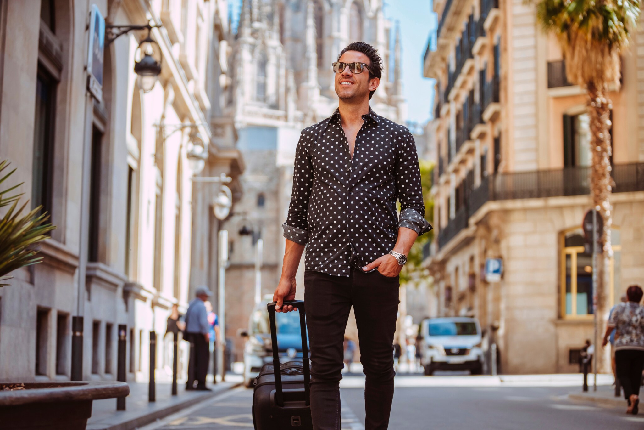 Ein elegant gekleideter Mann mit Sonnenbrille läuft mit einem schwarzen Trolley durch eine Straße in einer mediterranen Stadt.