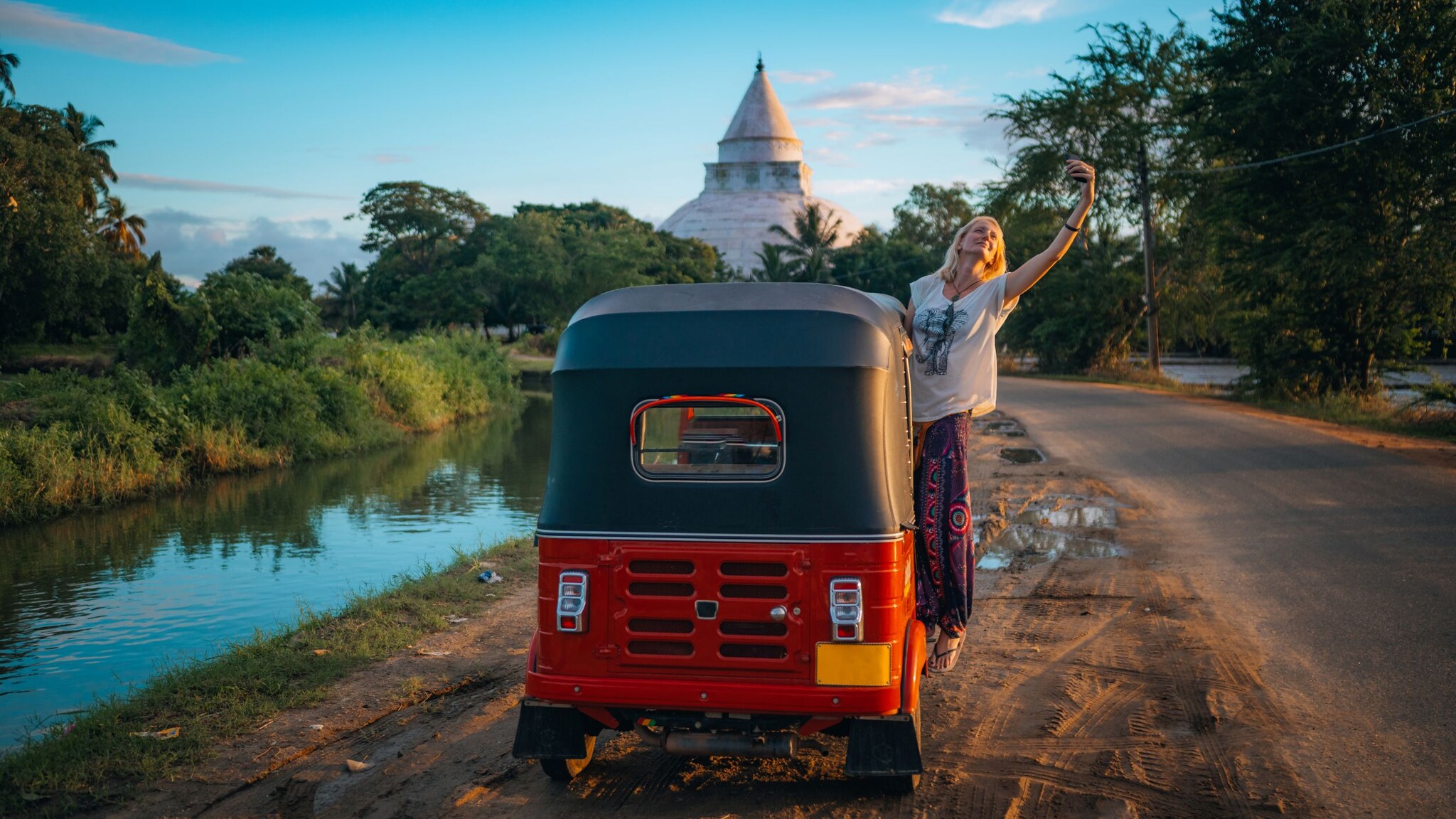 Frau macht Selfie neben rotem Tuk-Tuk auf ländlichem Weg mit Tempel im Hintergrund.