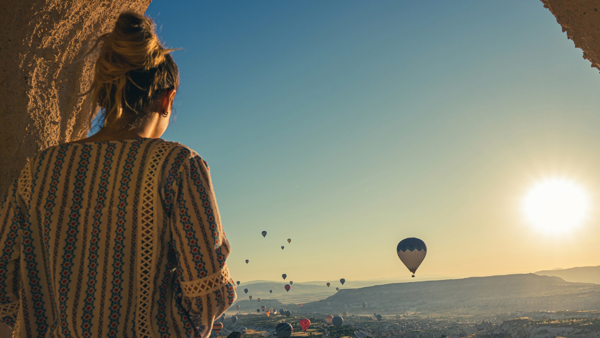 Eine Person mit einem langen, gemusterten Oberteil steht mit dem Rücken zur Kamera und blickt auf eine Landschaft mit vielen Heißluftballons im Himmel bei Sonnenaufgang.