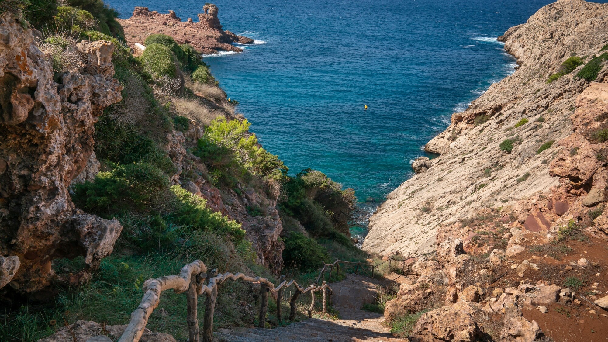 Steinerne Treppe mit Holzgeländer, die zu einem blauen Meer führt, umgeben von Felsen und grüner Vegetation. Steinerne Treppe mit Holzgeländer, die zu einem blauen Meer führt, umgeben von Felsen und grüner Vegetation.