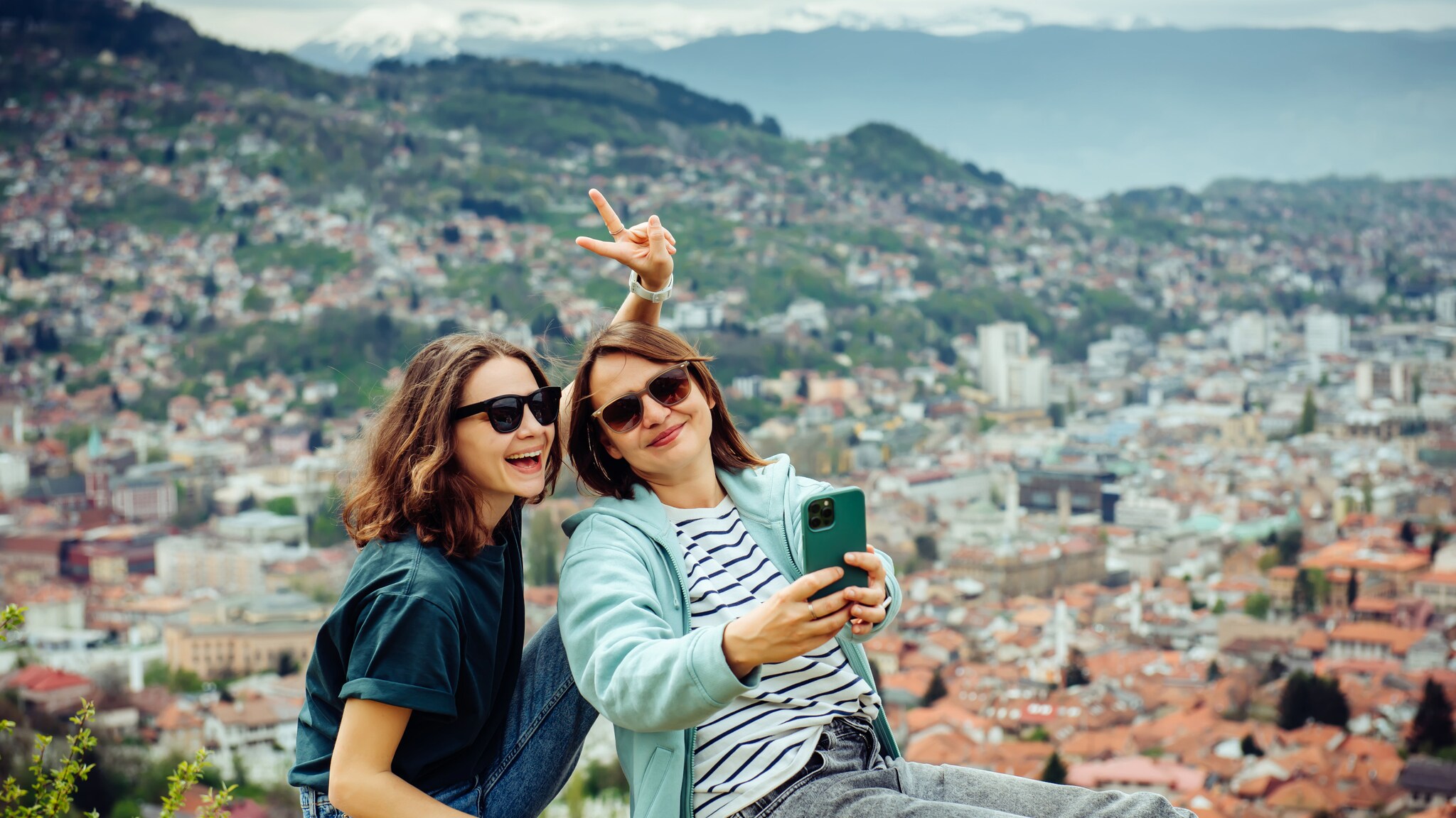 Zwei junge Frauen sitzen auf einer Mauer mit Blick auf eine Stadt, eine macht ein Selfie mit einem Smartphone und zeigt ein Peace-Zeichen.