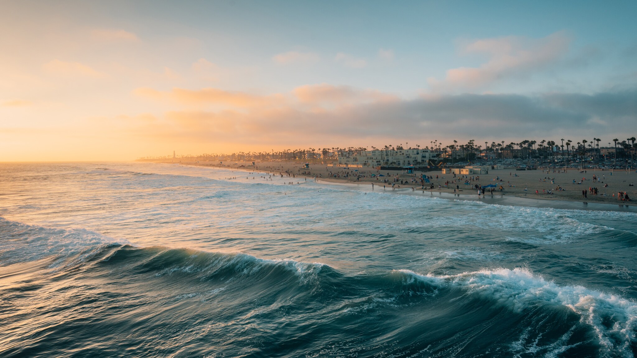 Sonnenuntergang über einem belebten Strand mit Wellen im Vordergrund und Palmen am Horizont