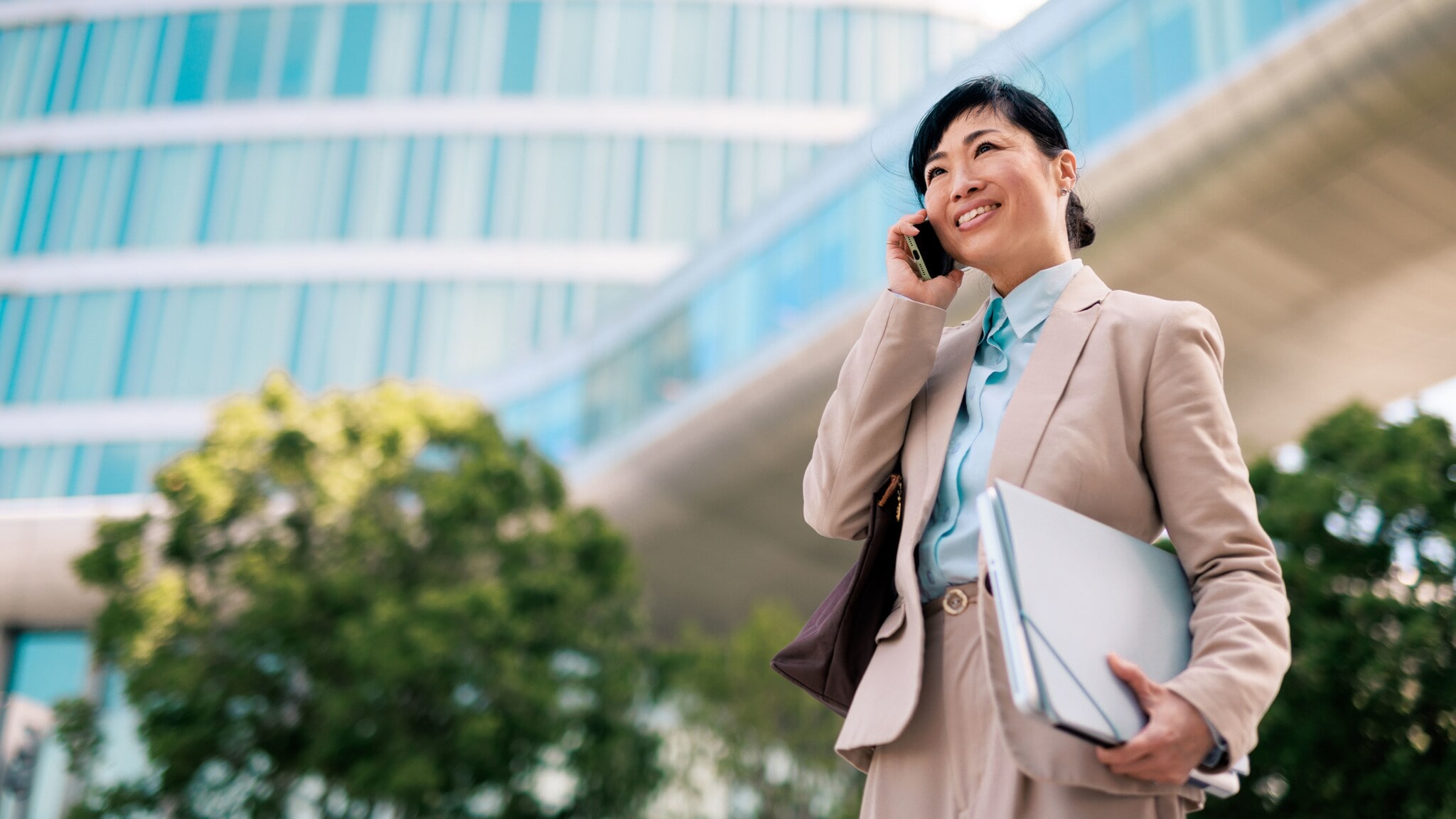 Eine lächelnde Frau im hellen Hosenanzug mit Laptop und Arbeitsmappe unterm Arm hält ein Smartphone ans Ohr vor einem großen Gebäude mit viel Glas. Eine lächelnde Frau im hellen Hosenanzug mit Laptop und Arbeitsmappe unterm Arm hält ein Smartphone ans Ohr vor einem großen Gebäude mit viel Glas.